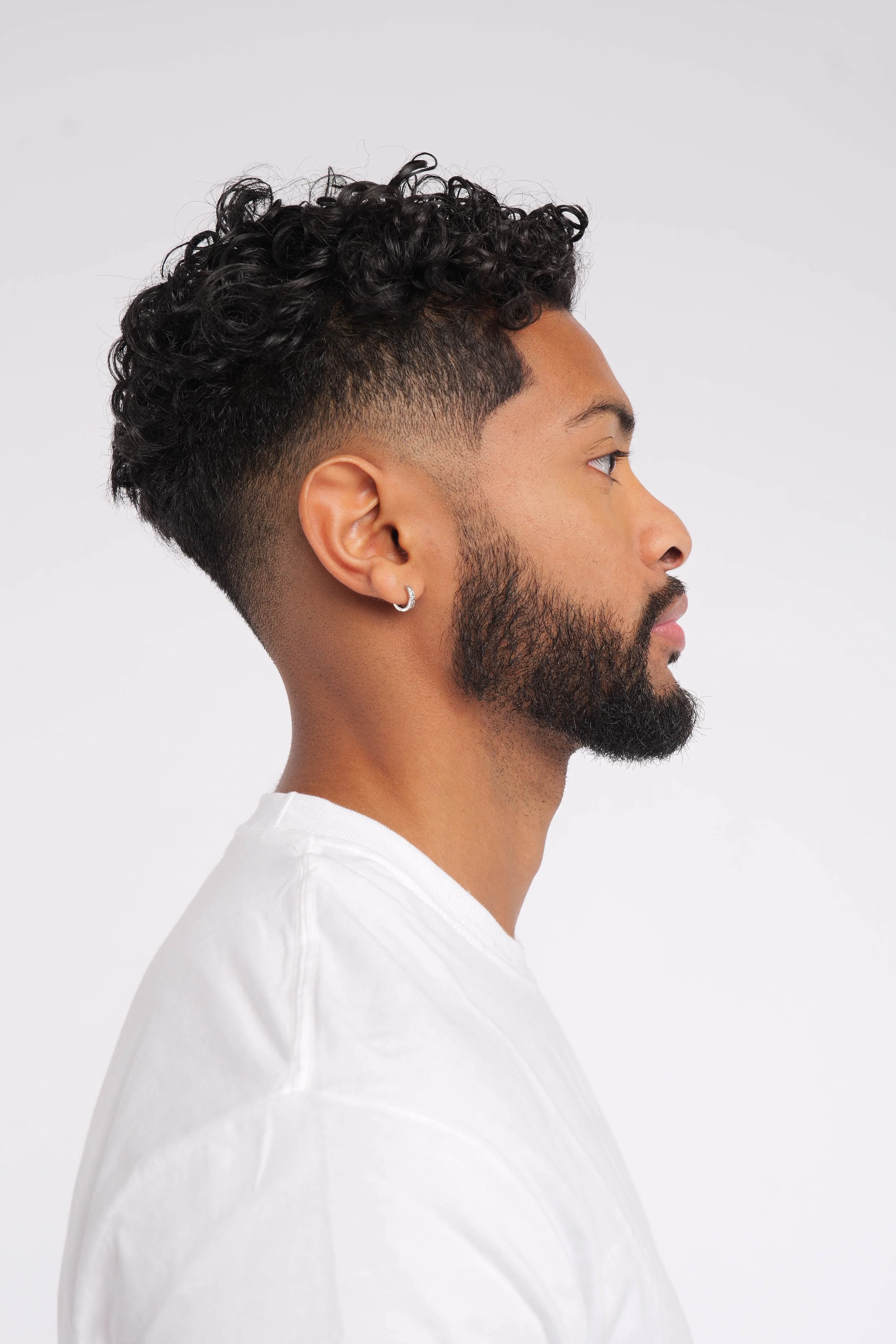 Profile of a man with a curly fade haircut, beard, and small hoop earring, wearing a white shirt.