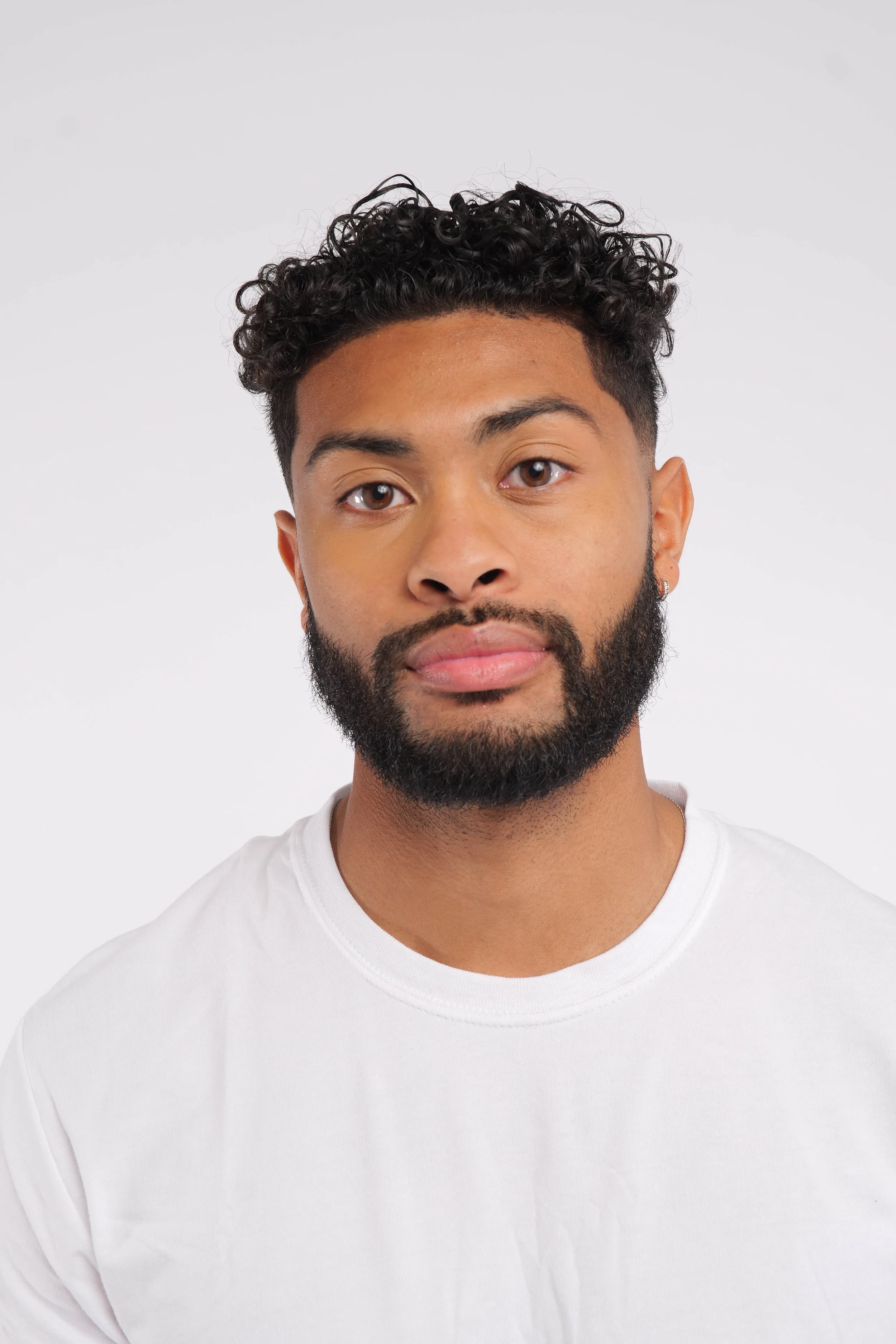 Headshot of a young man with curly black hair and a beard, wearing a white t-shirt, against a plain white background.