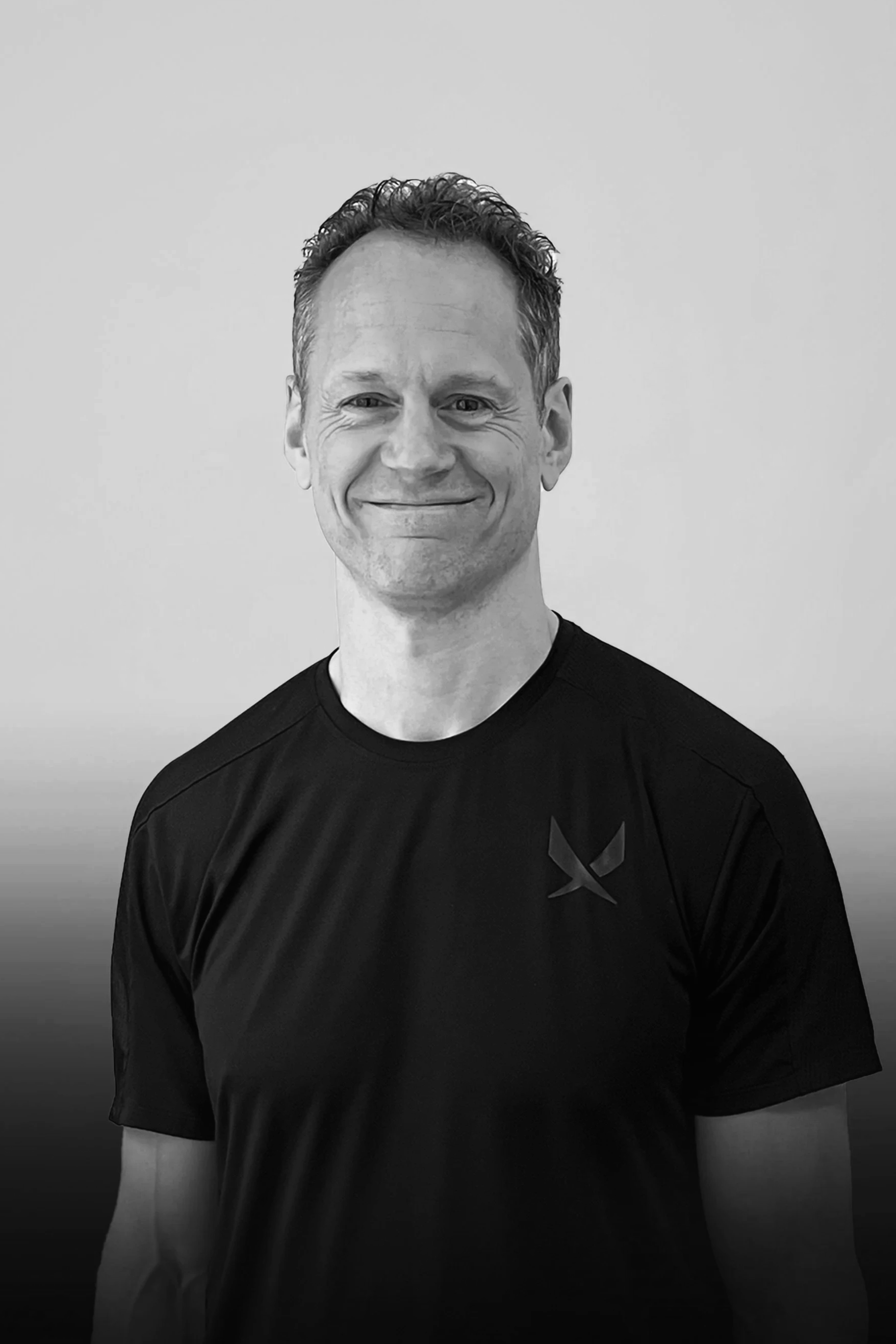 A smiling man with short, curly hair wearing a black athletic shirt with a small logo on the chest, standing against a plain light-colored background.