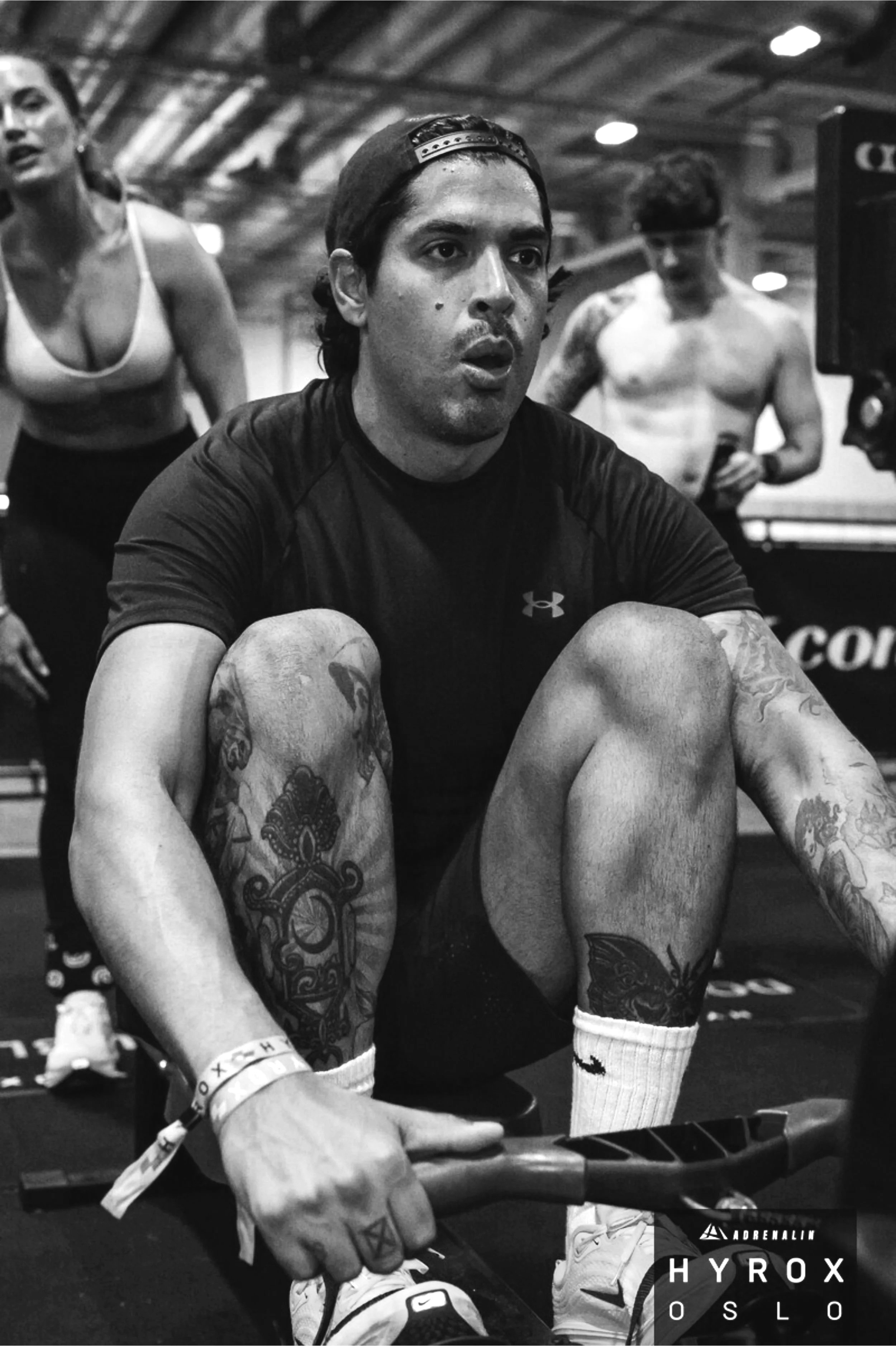 A man with tattoos on his arms and legs, wearing a black athletic shirt, sitting on an exercise bike in a gym, with two other people in workout clothes in the background.