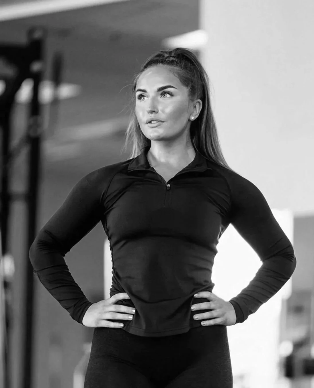 A woman in black athletic clothing standing with hands on her hips in an indoor gym or fitness studio.