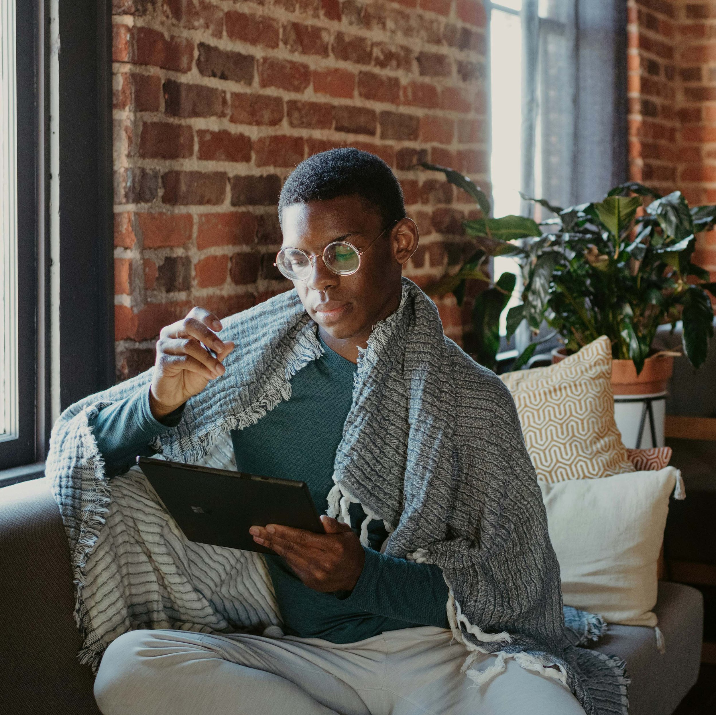 A person sitting on a couch, wearing glasses, reading on a tablet, with a blanket draped over their shoulders, next to a window and a large plant.