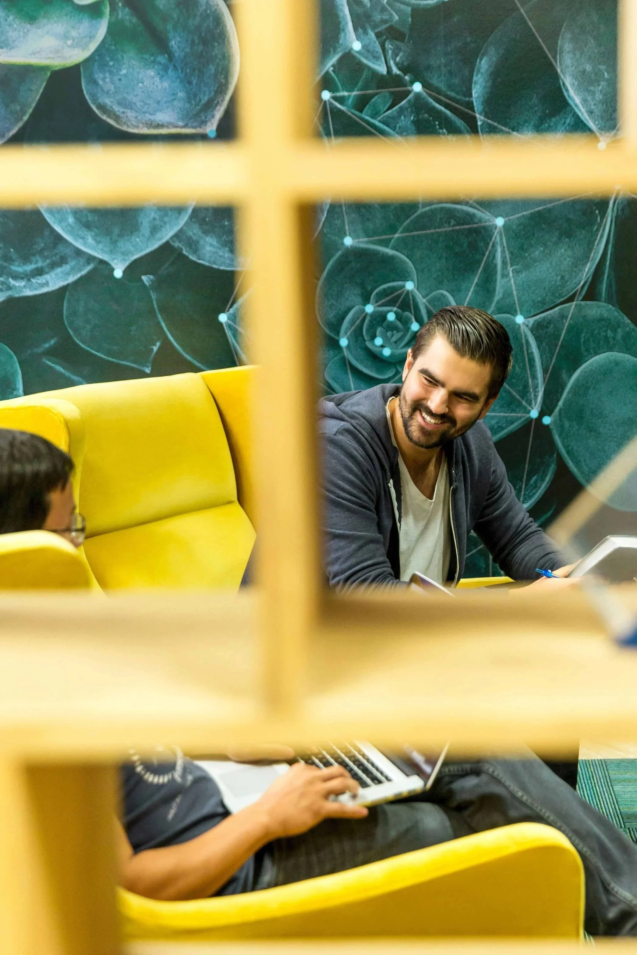 Two men sitting on yellow couches in a room with a leafy green and molecular patterned wall, seen through a window with yellow framing, engaging in a conversation with one man smiling.