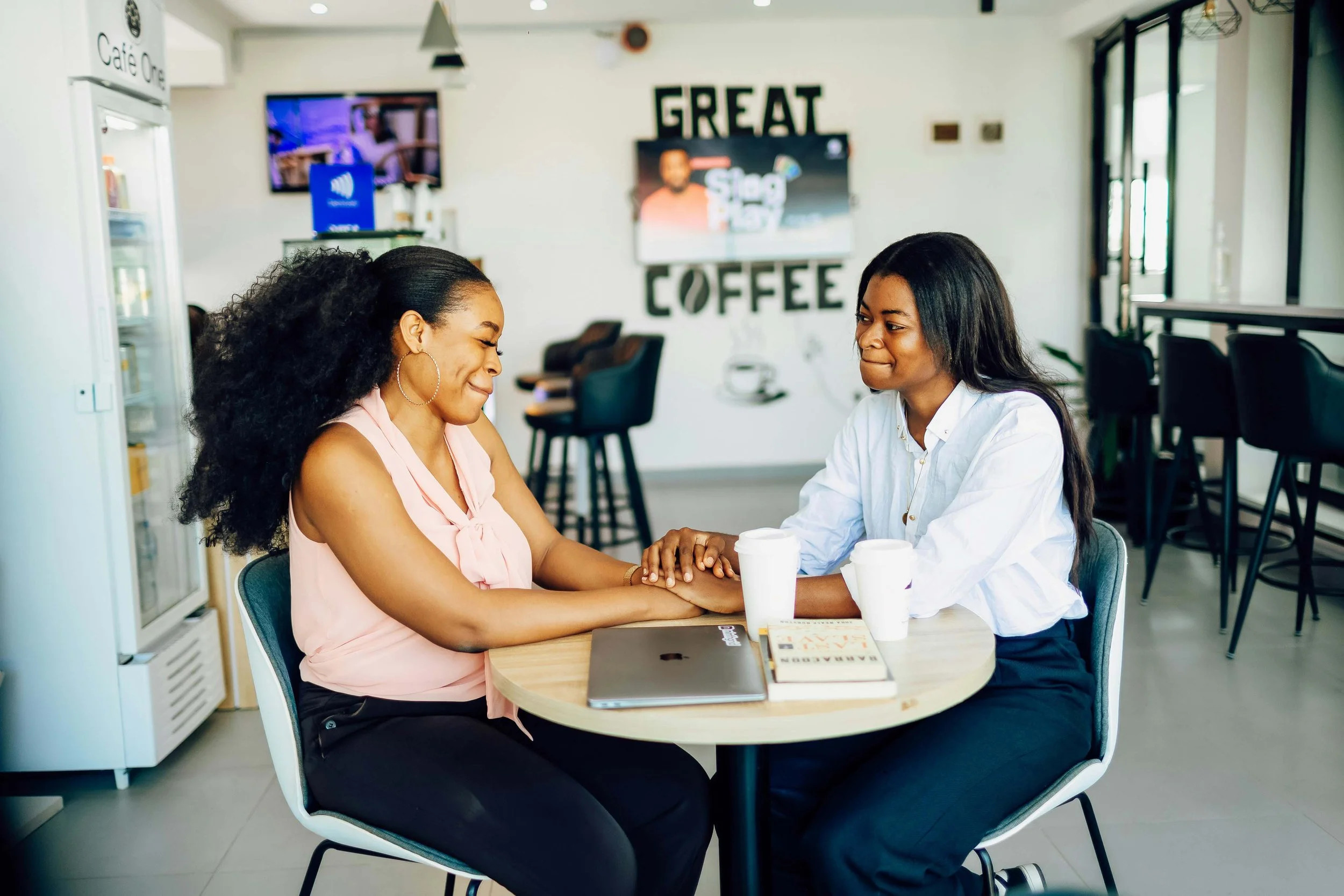 Two women sitting at a table in a coffee shop, holding hands and smiling at each other, with coffee cups and a laptop on the table.