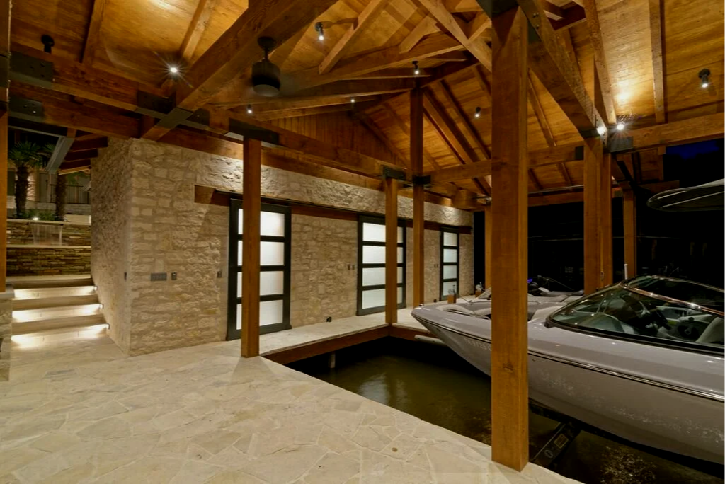 Interior of a luxurious boathouse featuring wooden beams, stone walls, frosted glass double doors, and a boat docked in water.