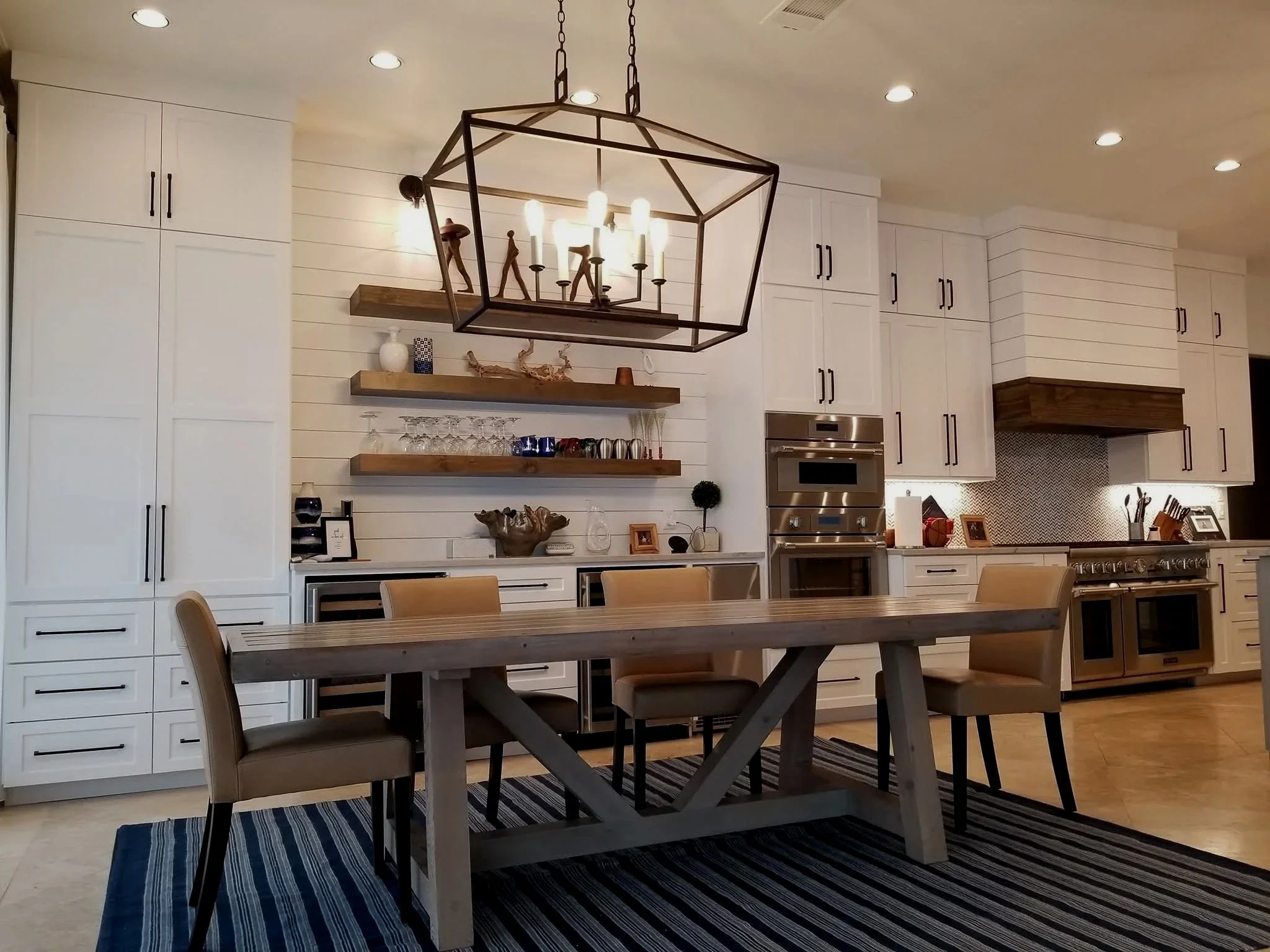 Modern kitchen with white cabinets, stainless steel appliances, a wooden dining table with beige chairs, a striped rug, and decorative floating shelves.