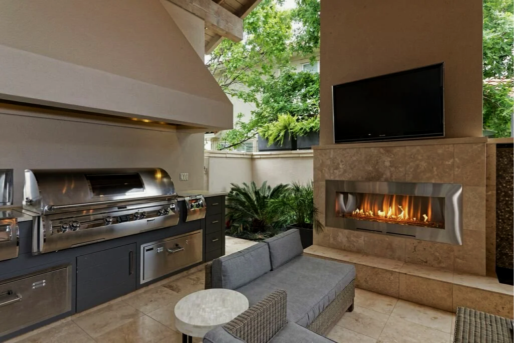Outdoor living area with a built-in grill, a mounted flat-screen TV, a modern fireplace, a gray outdoor sofa, and lush greenery outside.