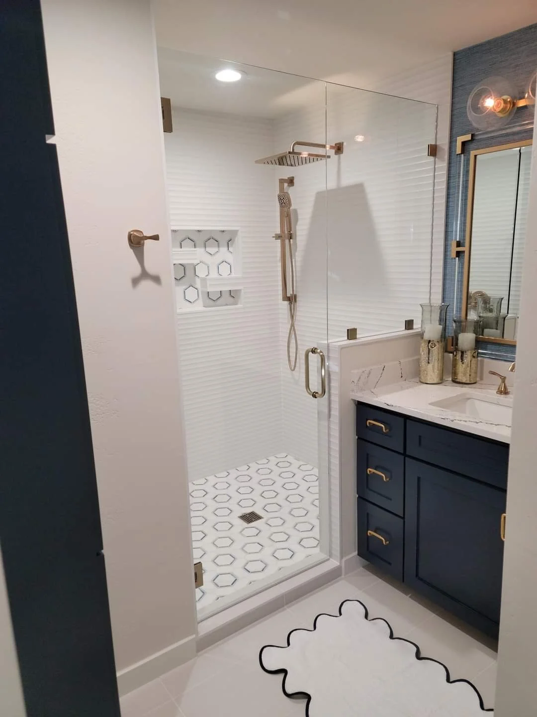 A modern bathroom shower area with clear glass door, rain shower head, and a built-in wall niche with decorative tiles. Adjacent to the shower is a navy blue vanity with a white marble countertop, gold fixtures, and decorative candle holders.