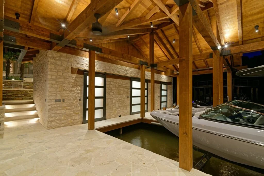 Interior of a luxurious boathouse featuring wooden beams, stone walls, frosted glass double doors, and a boat docked in water.