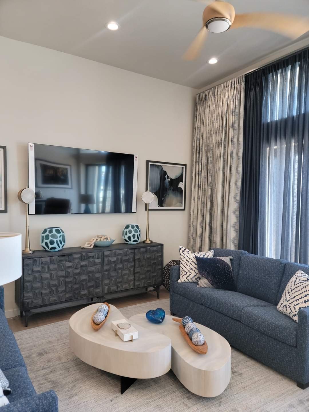 Living room with a dark grey sofa, modern light wood coffee table with decorative objects, a black textured sideboard under a mounted flat-screen TV, framed black and white artwork, a white and blue ceramic lamp, beige and navy curtains, and ceiling 