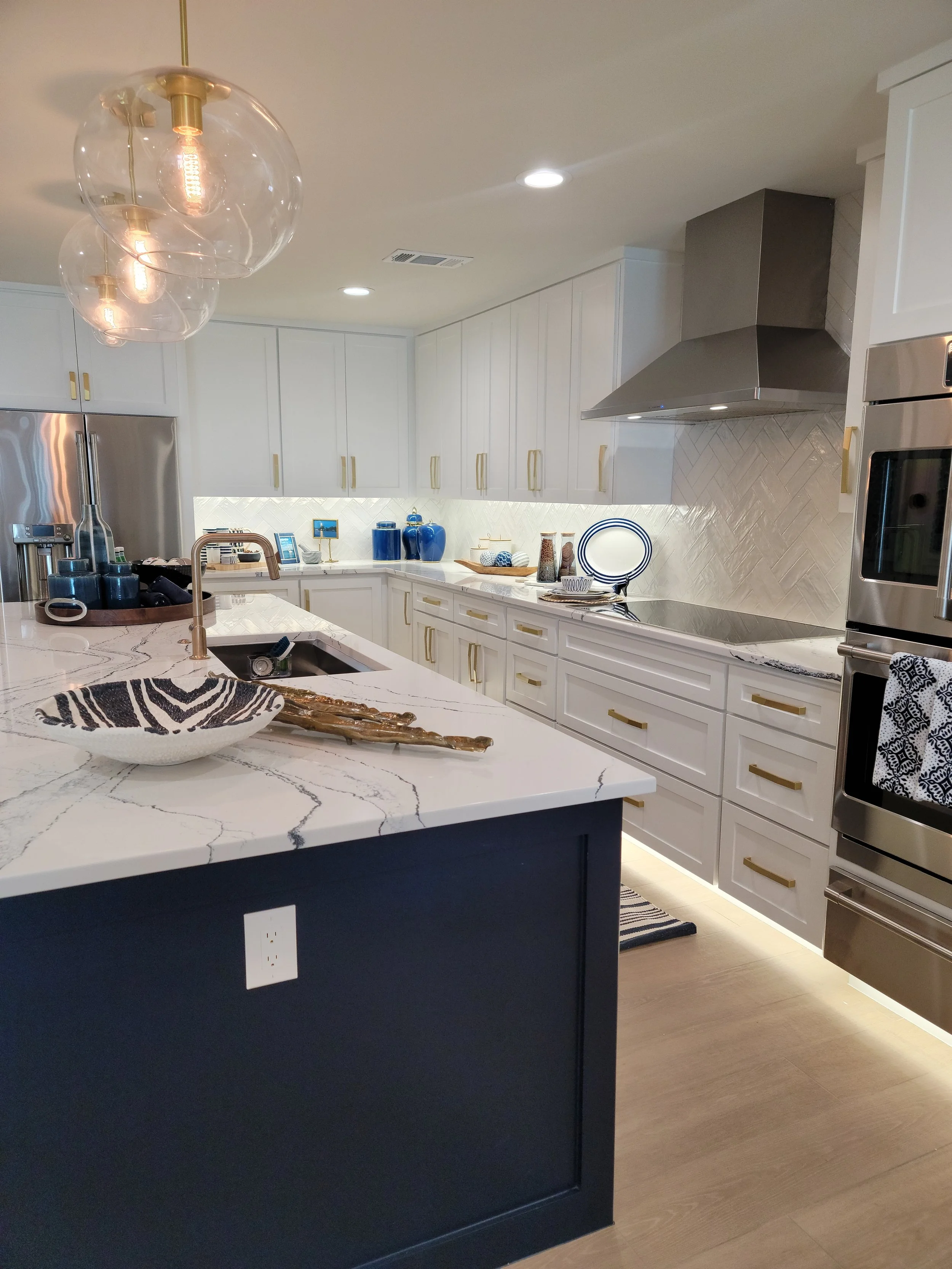 Modern kitchen with white cabinets, marble countertops, gold handles, stainless steel appliances, and decorative blue accents. Overhead lighting fixtures and under-cabinet lighting illuminate the space.