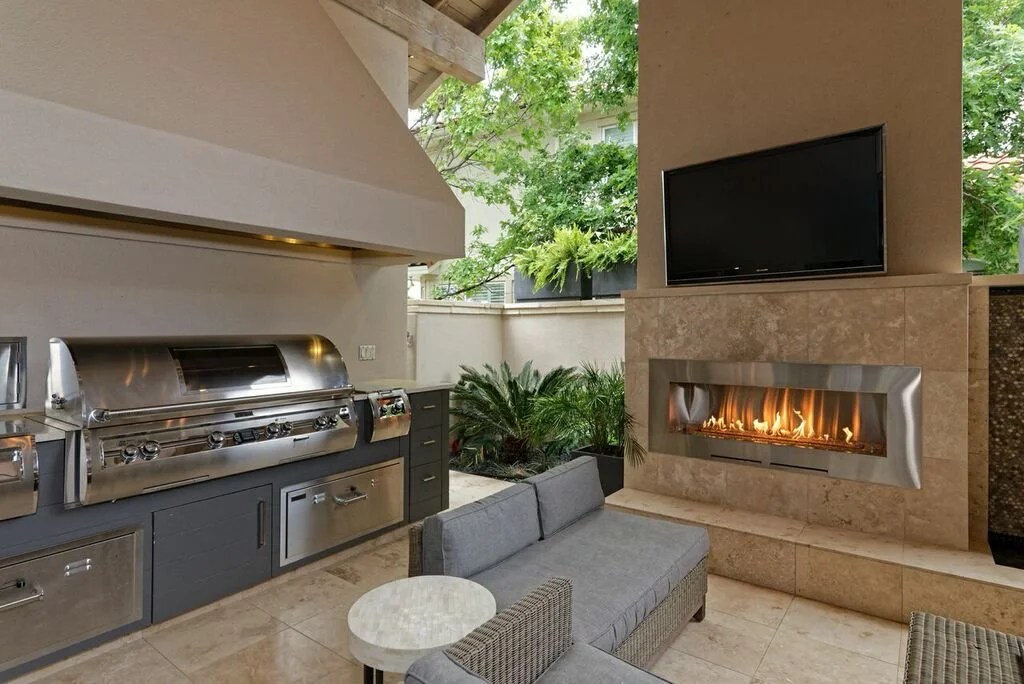 Outdoor living area with a built-in grill, a mounted flat-screen TV, a modern fireplace, a gray outdoor sofa, and lush greenery outside.