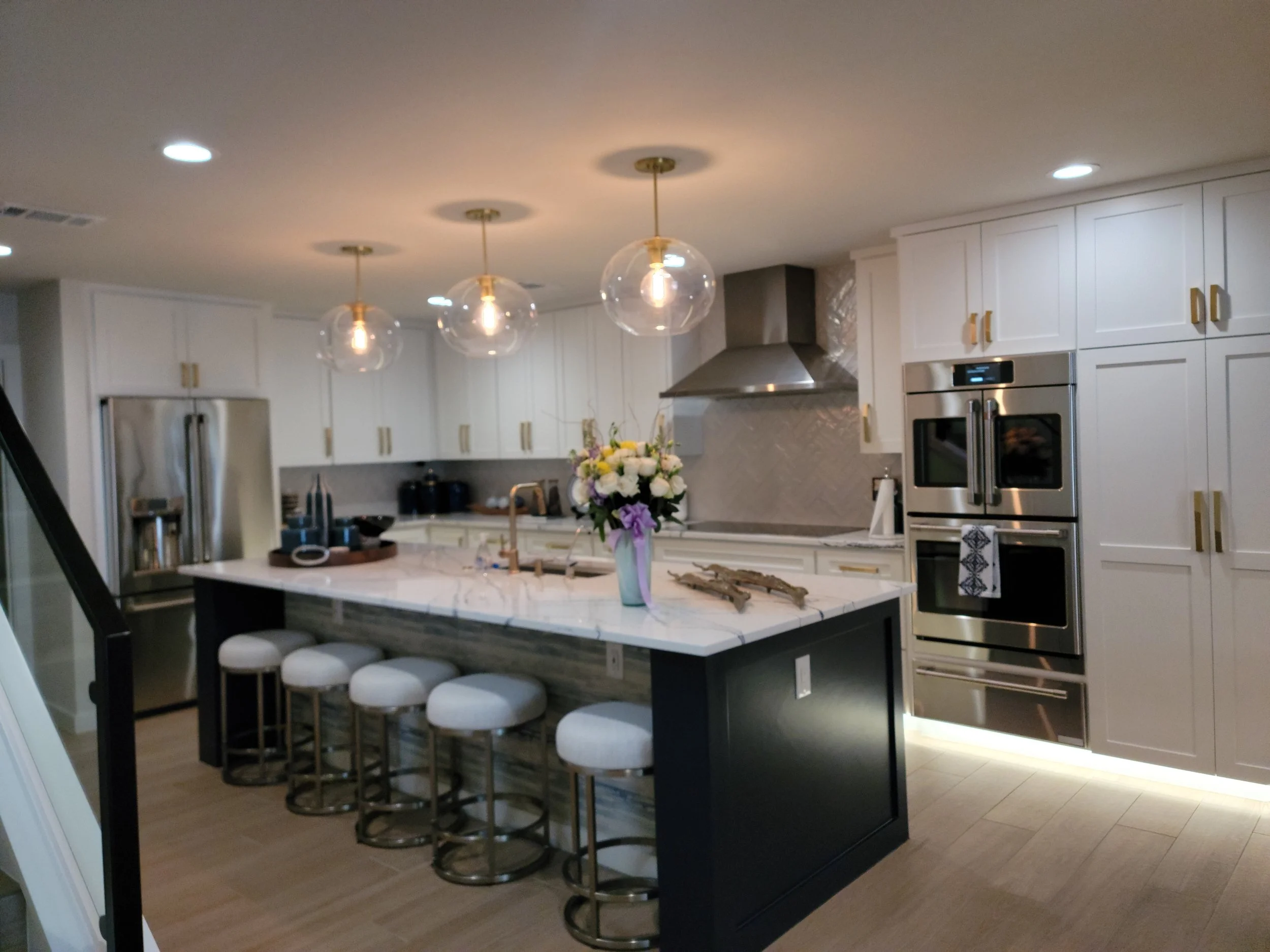 Modern kitchen with white cabinets, stainless steel appliances, a central island with a marble countertop, and four white cushioned barstools. Hanging from the ceiling are three glass pendant lights. The island features a vase with flowers and decora