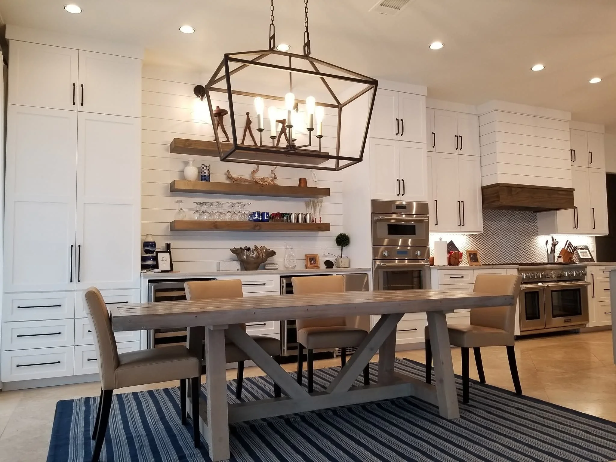 Modern kitchen with white cabinets, stainless steel appliances, a wooden dining table with beige chairs, a striped rug, and decorative floating shelves.