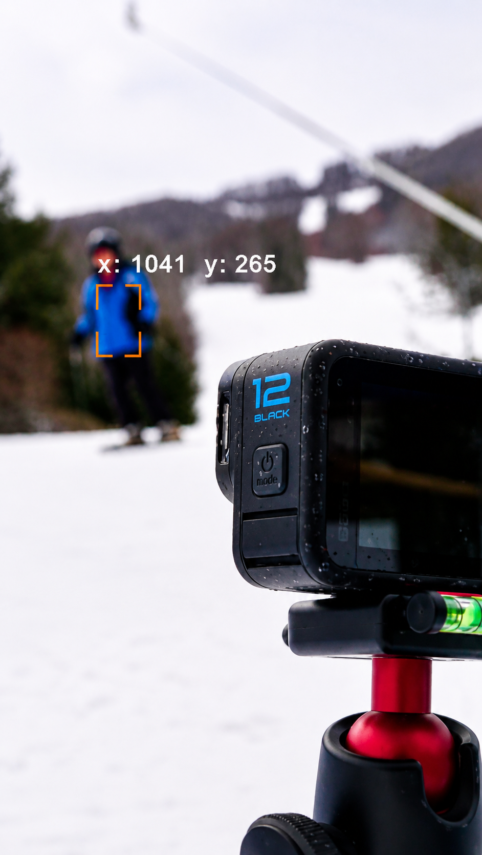 A digital camera on a tripod capturing a person in a blue jacket and black pants snowboarding on a snowy slope. The background shows snow-covered mountains and trees, with an out-of-focus skier in the distance.