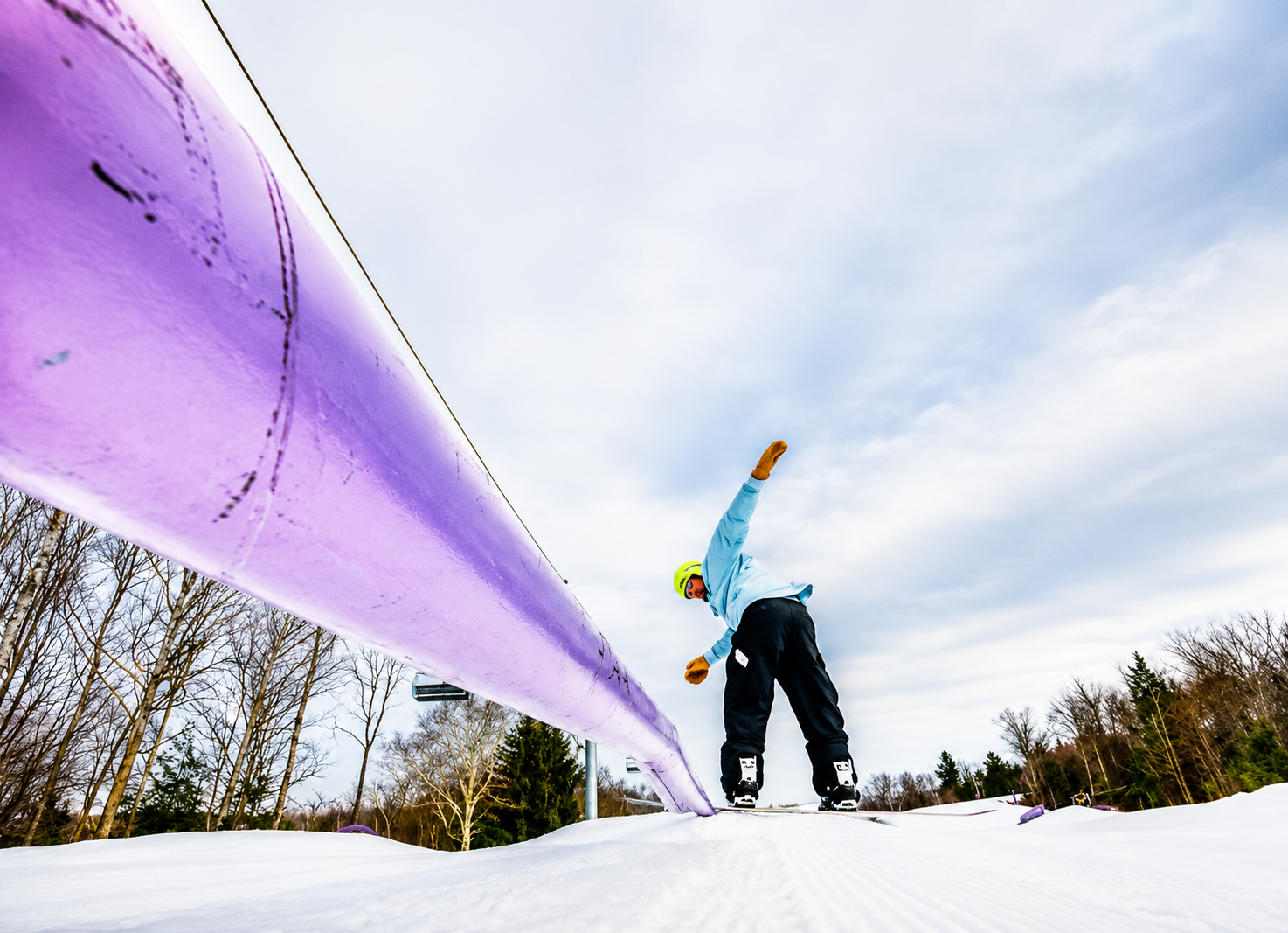 A person wearing a yellow helmet, winter gloves, and a light blue jacket is leaning backward while snowboarding on a snowy slope. A large purple snow rail is in the foreground. Trees and a cloudy sky are in the background.