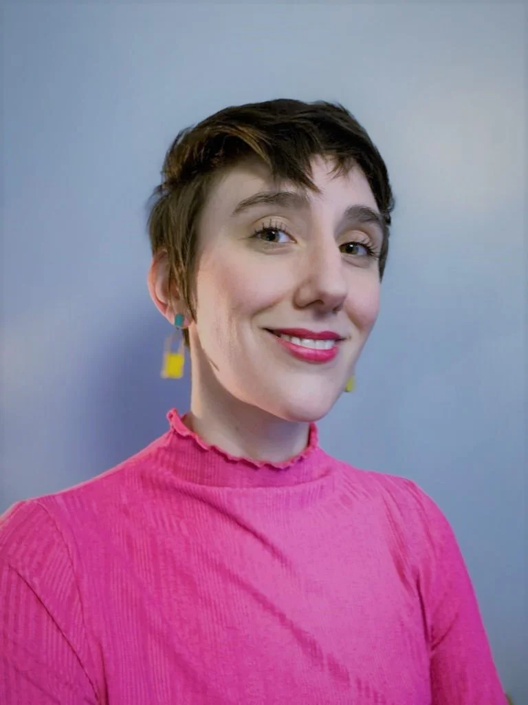 A young woman with short brown hair wearing a pink turtleneck top and colorful earrings, smiling at the camera against a plain blue background.
