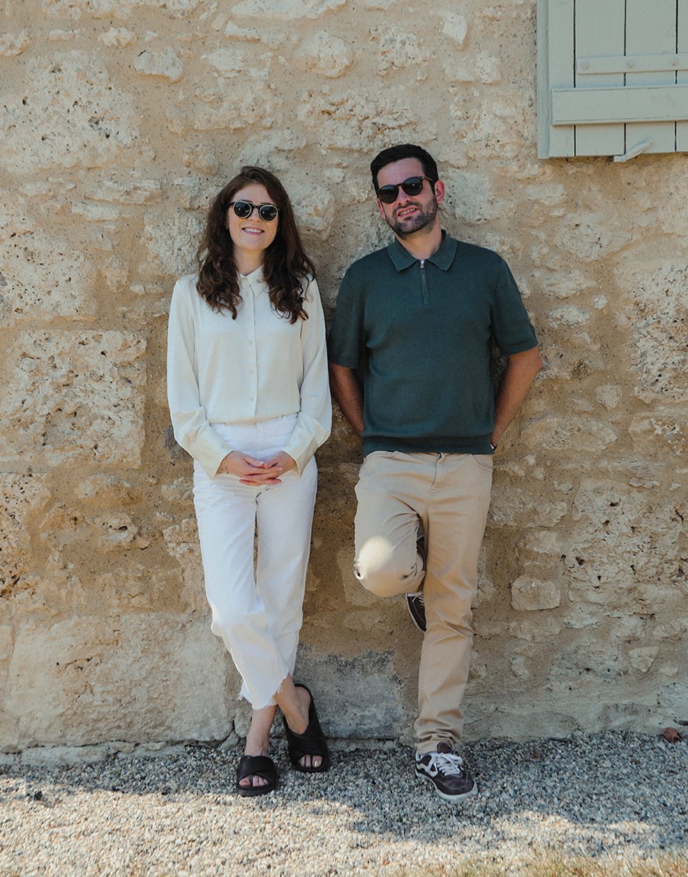 A man and woman standing against a beige stone wall, both wearing sunglasses. The woman is dressed in white with black sandals, and the man is wearing a dark green shirt, beige pants, and sneakers.