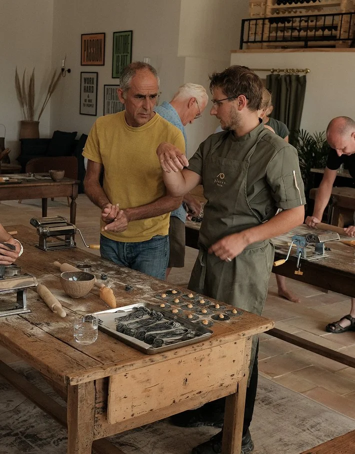 People participating in a pasta-making class, one person receiving a hand massage from the instructor as another observes, with pasta tools and ingredients on a rustic wooden table.