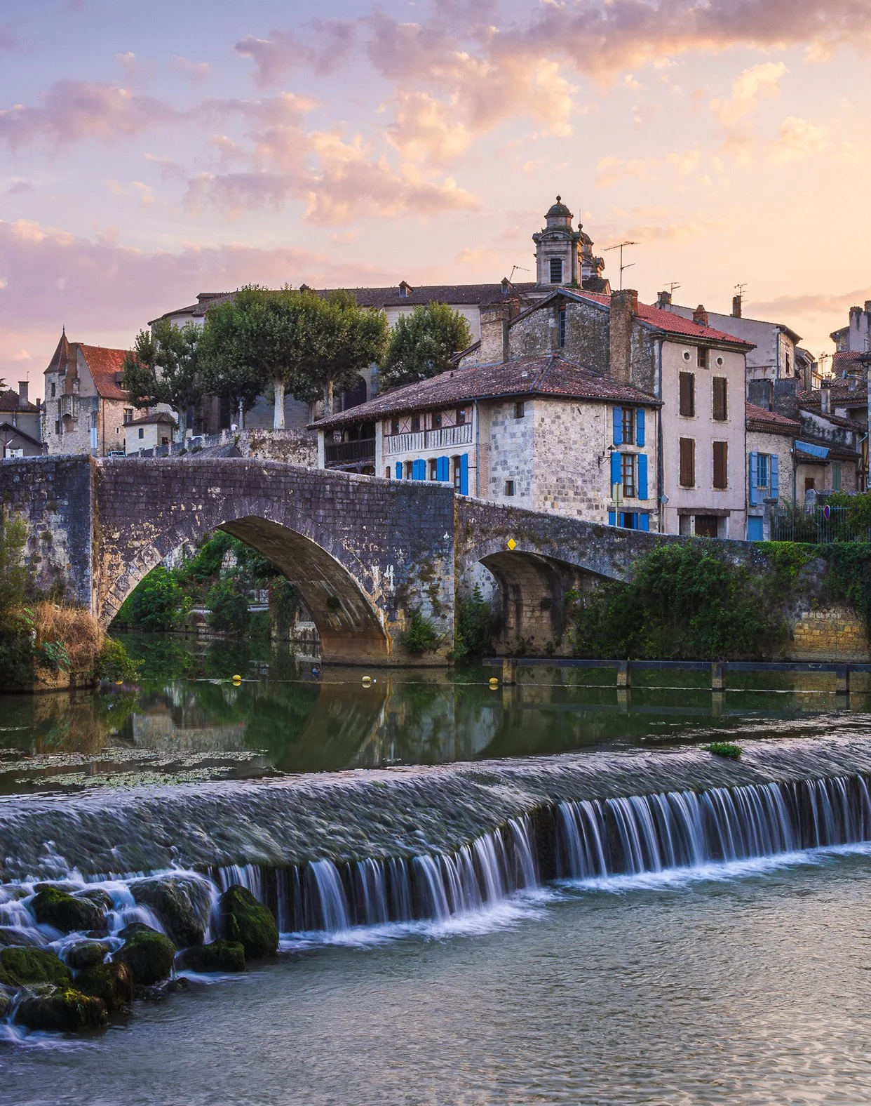 Scenic view of a historic stone bridge over a river with a small waterfall in a European town at sunset, with old buildings and trees in the background.