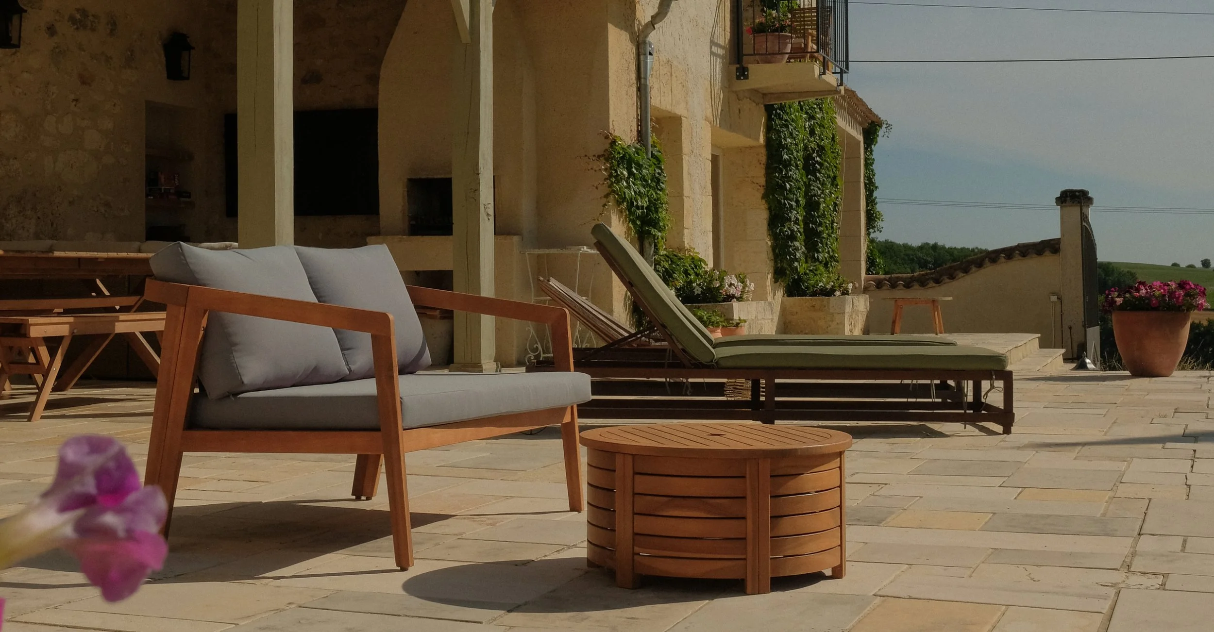 A patio with lounge chairs, a cushioned wooden couch, a small round wooden side table, potted plants, and a scenic background of fields and plants under a clear sky.