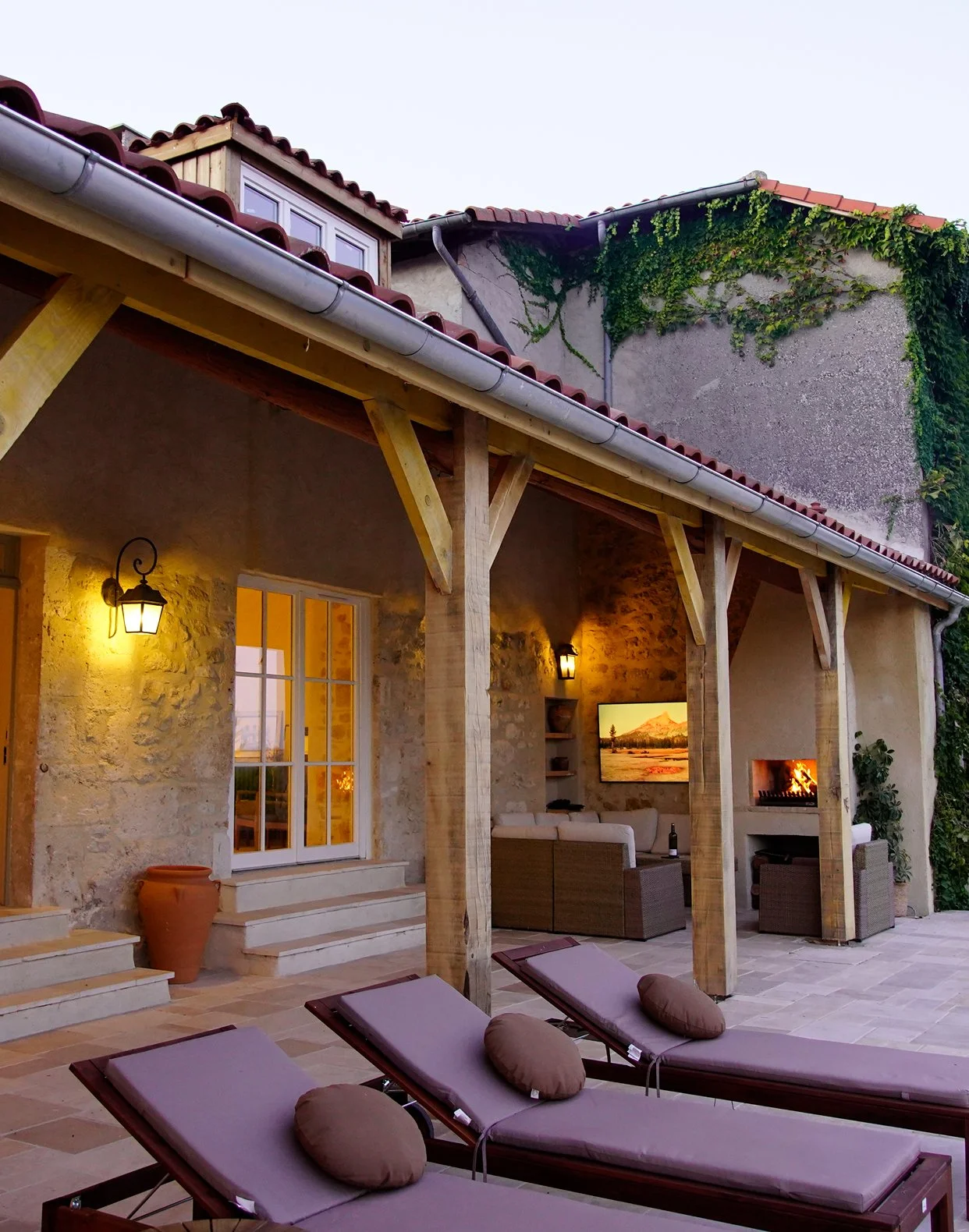 Outdoor patio area of a house with three lounge chairs, a fireplace, and a TV, with a stone wall exterior and ivy growing on upper wall. Warm lighting in the evening.