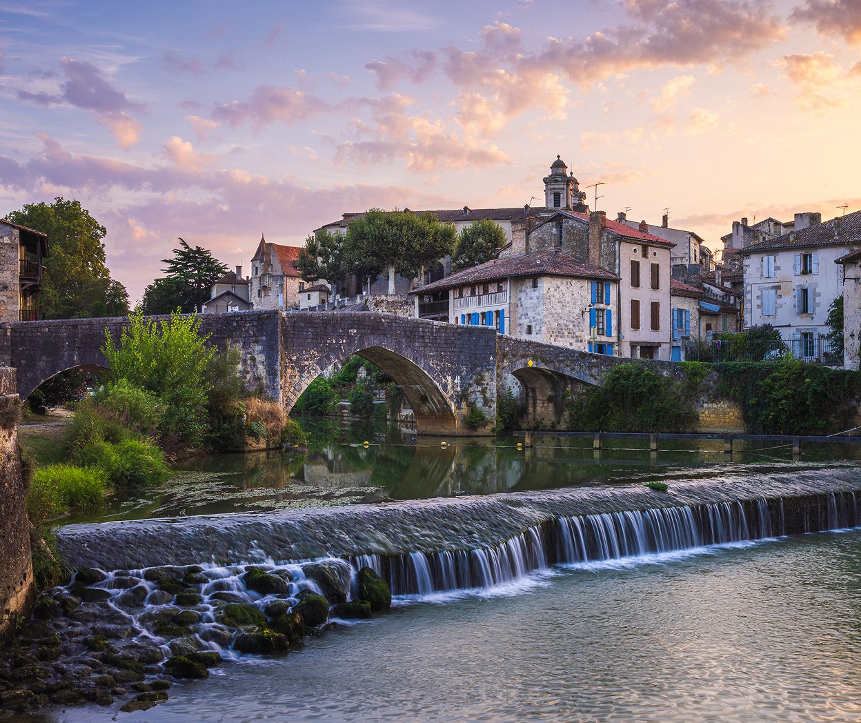 A scenic view of a small European town with a stone arched bridge over a river, medieval buildings, and a church tower in the background under a partly cloudy sky at sunset.