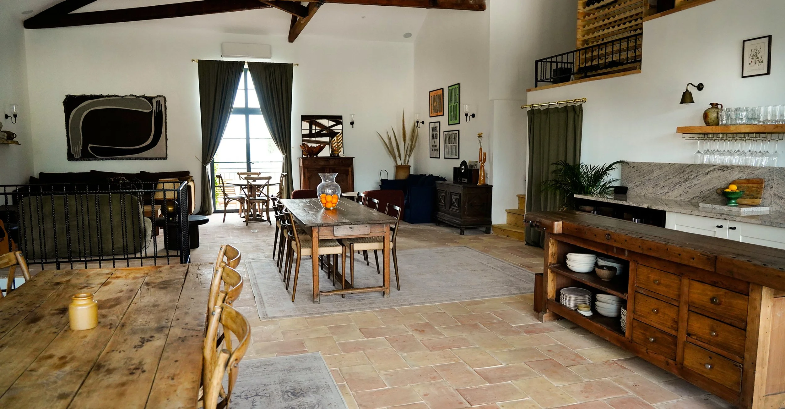 Spacious open-concept living and dining area with wooden furniture, a large window with green curtains, and art on the walls. There is a wooden kitchen island with dishes and a marble countertop, along with various decorative items and plants.