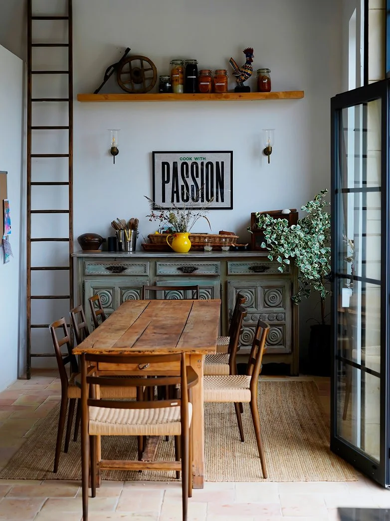 A cozy dining area featuring a wooden table with six chairs. The background includes a vintage sideboard with a yellow vase, dried flowers, and kitchen utensils. Wall decor includes a framed sign saying 'Cook with Passion' and two wall-mounted lights. A shelf above holds jars, a wheel, and a decorative rooster. A plant is next to the sliding glass door on the right.