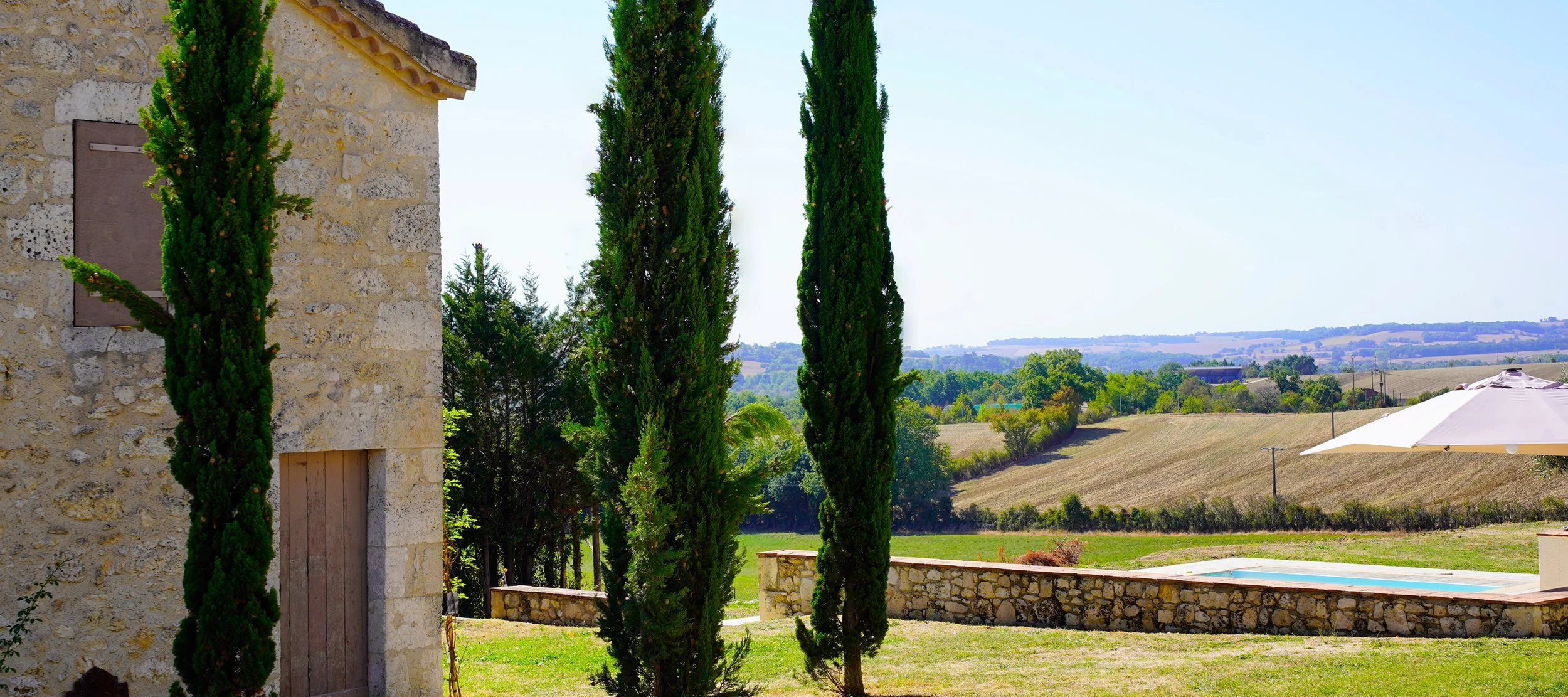 Stone cottage with wooden window shutters, three tall cypress trees, a stone wall, an umbrella, and rolling countryside in the background.