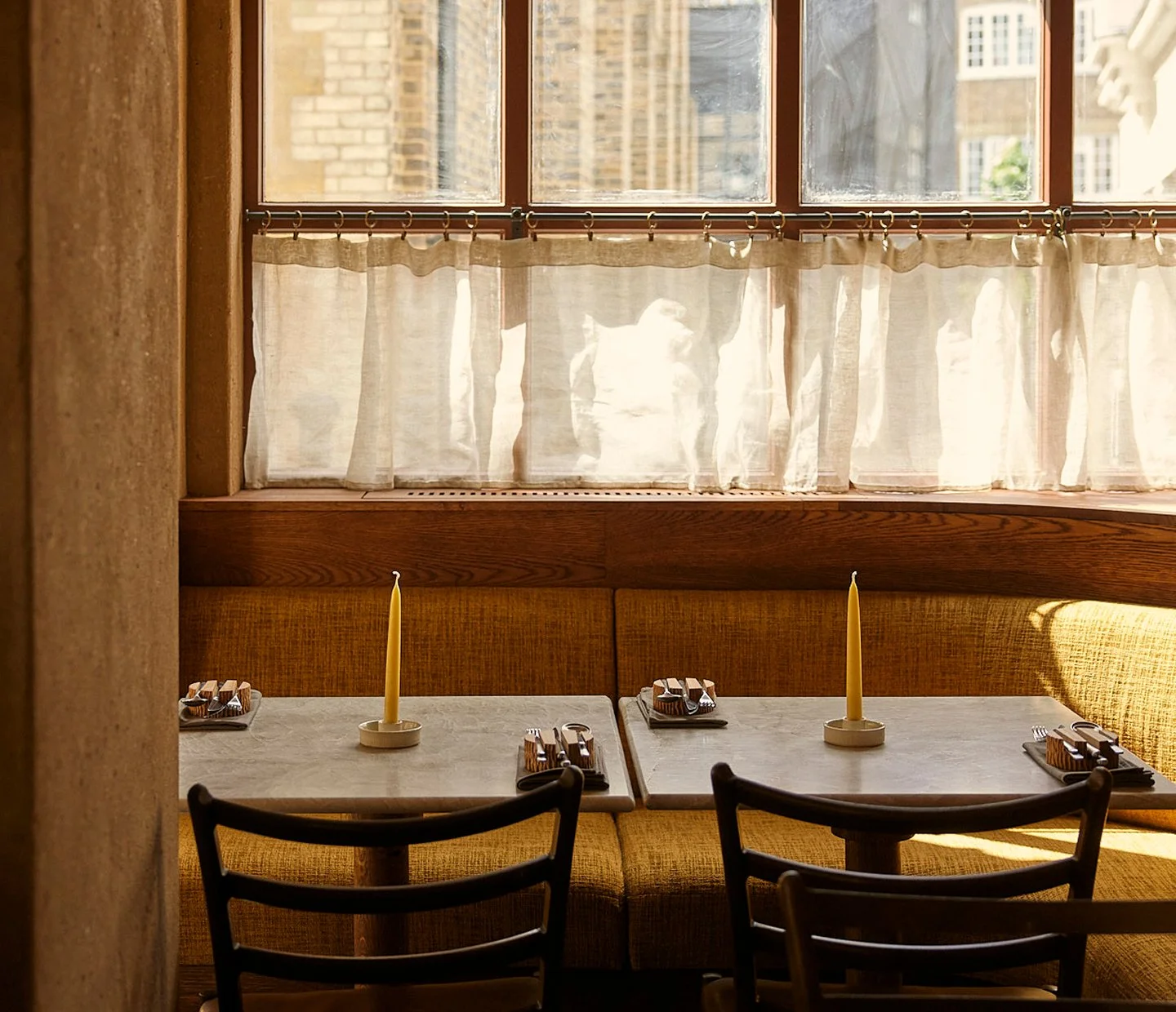 A cozy restaurant corner with two tables, each set with cutlery and a yellow candle. Sunlight streams through a large window with beige curtains, illuminating the yellow upholstered bench seating along the wall and creating a warm, inviting atmosphere.