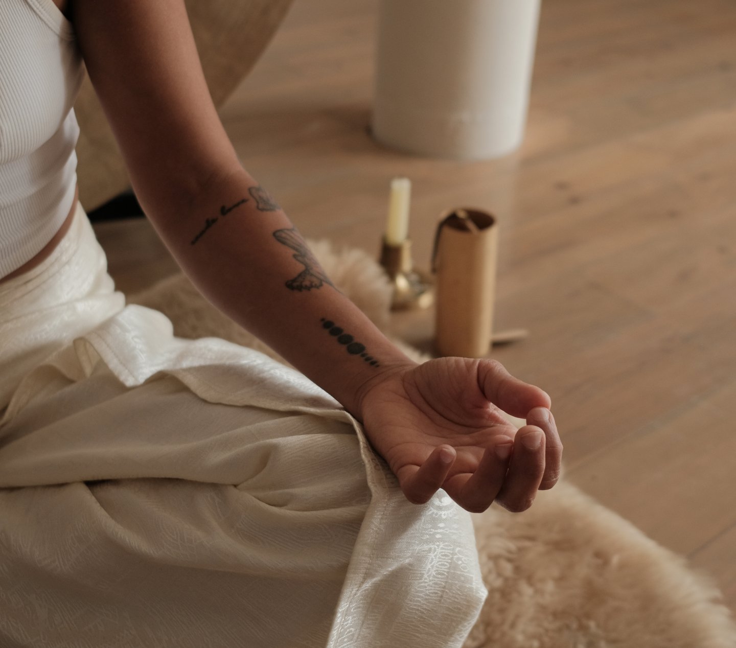 Person sitting cross-legged on a soft rug, wearing a white outfit, with tattoos on their forearm. In the background, there are candles and a cylindrical object on the floor, with a wooden floor and a white cylindrical piece of furniture visible.
