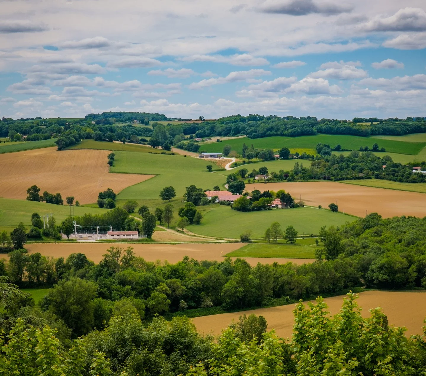 Rolling green hills with patches of farmland, trees, and a few buildings under a partly cloudy sky.