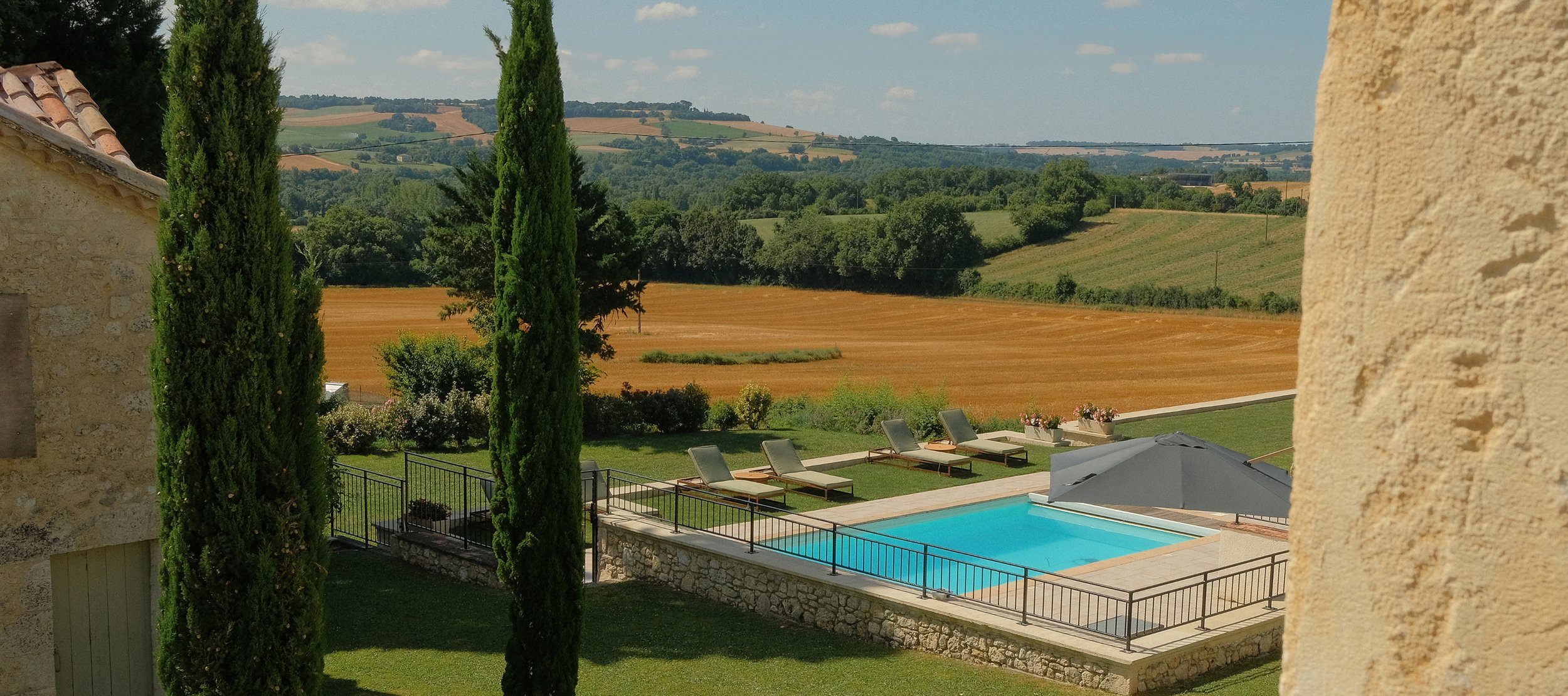 View of a swimming pool with lounge chairs, an umbrella, and a garden, overlooking rolling countryside with fields and trees under a partly cloudy sky.