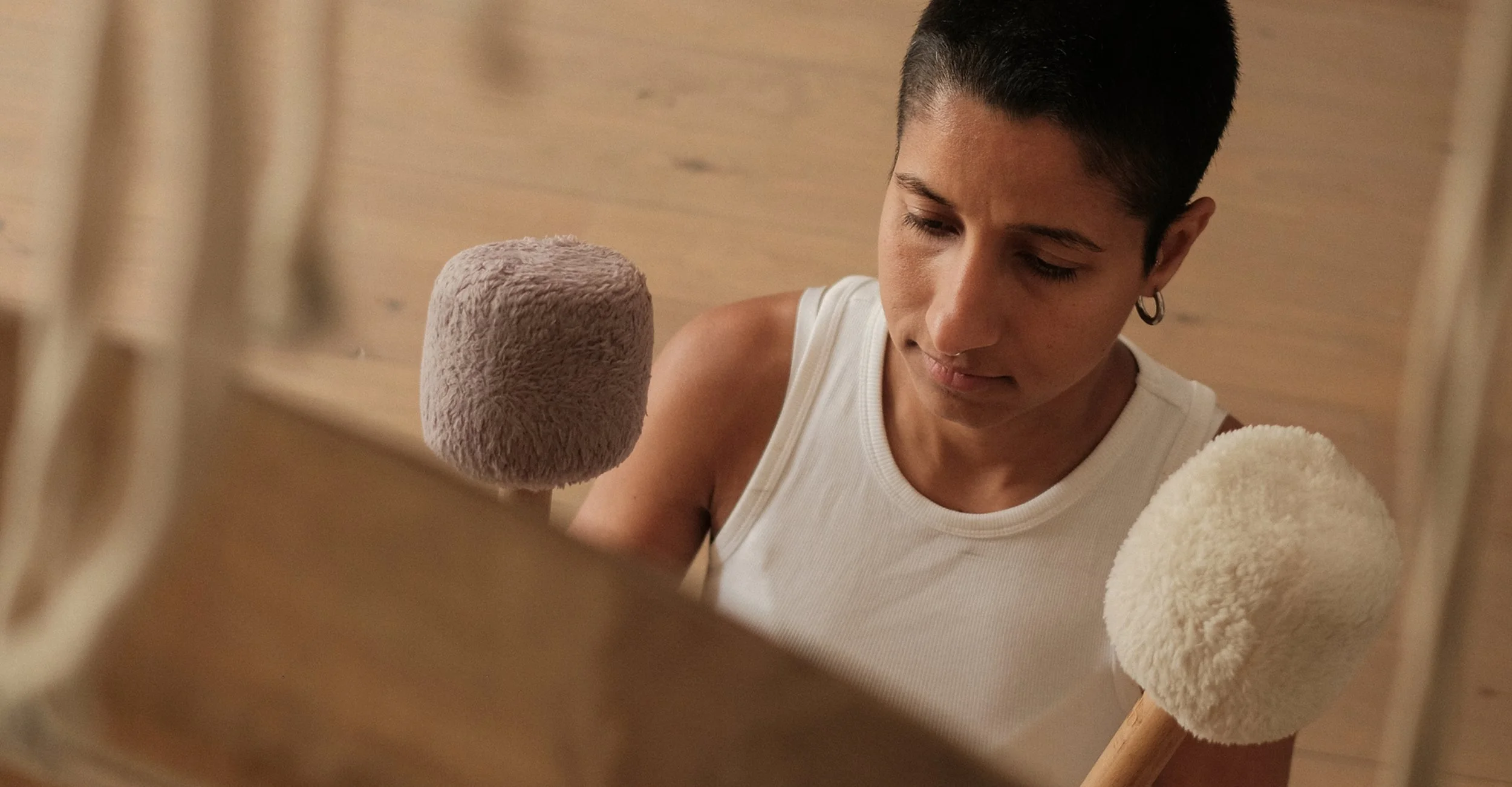 A person with short dark hair, wearing a white sleeveless shirt, is looking down at two fluffy mallet-like objects.
