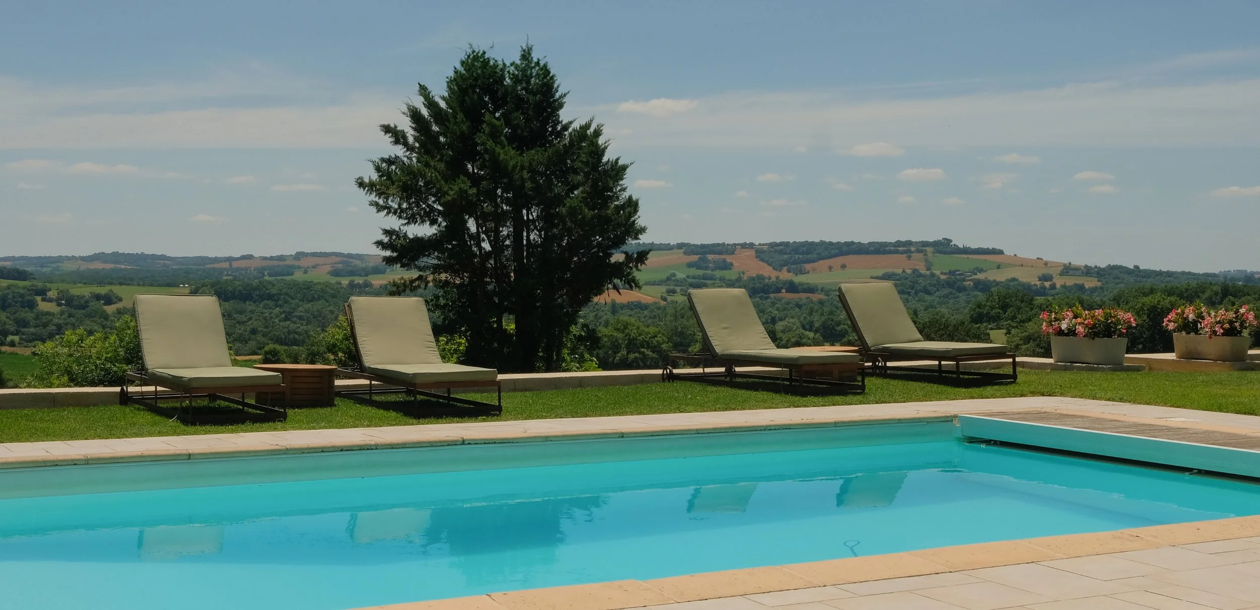 An outdoor swimming pool with four lounge chairs and a small side table on a grassy area, overlooking a scenic landscape of rolling hills under a blue sky with scattered clouds.