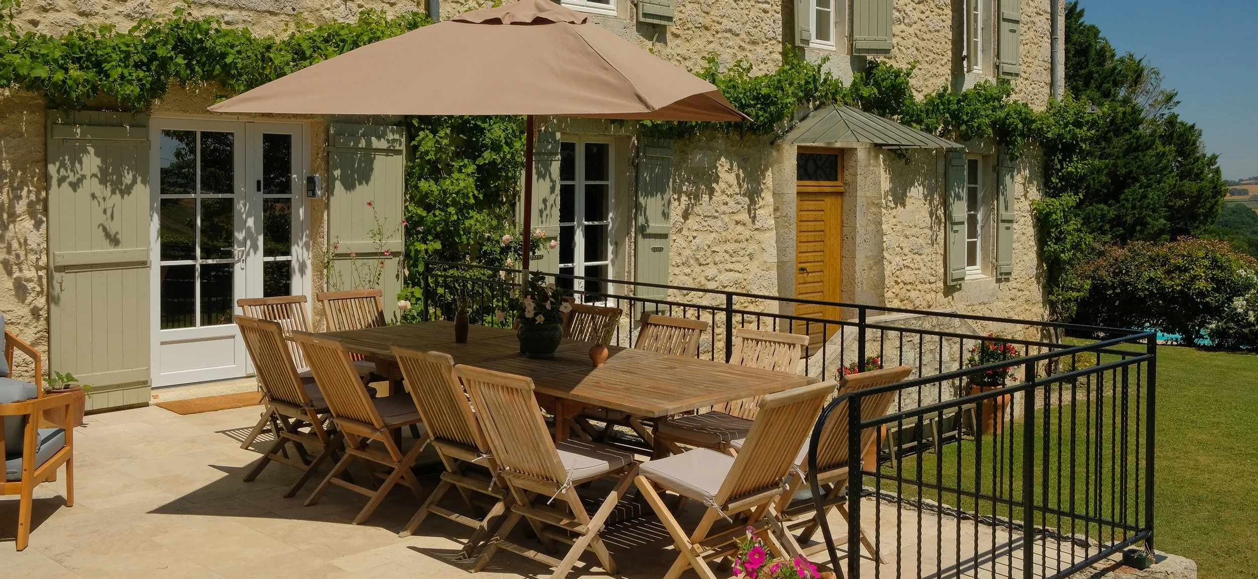 Outdoor patio area with a large wooden dining table and twelve wooden chairs, topped with cushions and an umbrella, adjacent to a stone house with green shutters, surrounded by greenery and flowers.
