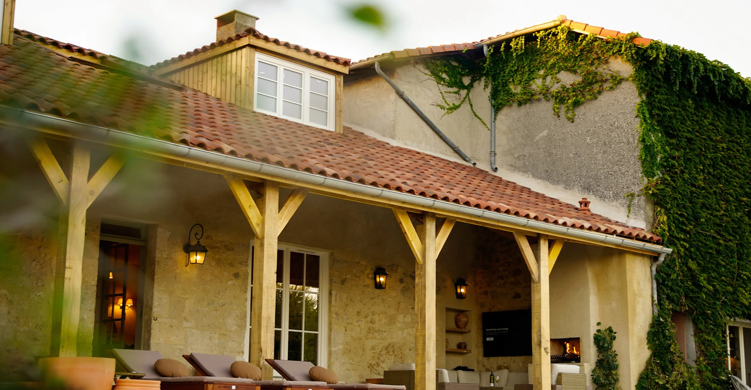 The image shows a rustic house with a beige stone exterior, a tiled roof, and a covered patio. The patio has lounge chairs, cushions, and outdoor lighting. Green ivy vines grow on the upper walls and roof area. There is a small chimney and a television on the patio wall.