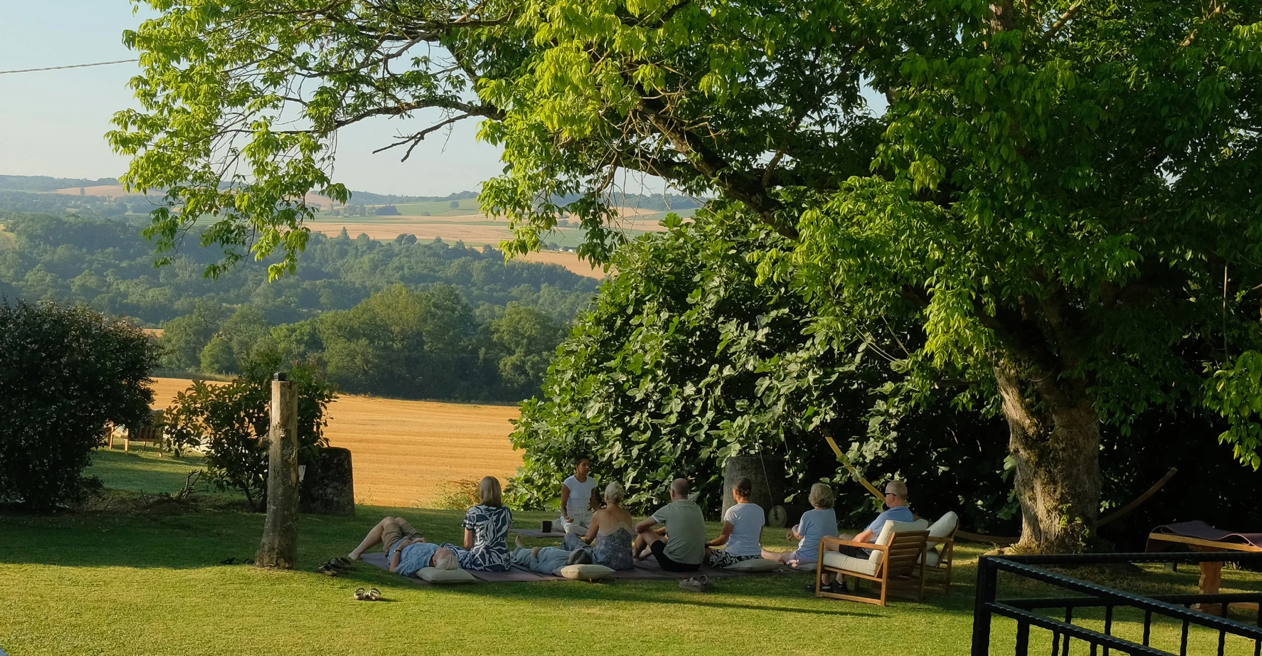 Group of people sitting on the grass and on chairs under a large tree, enjoying a peaceful outdoor gathering with a scenic view of rolling hills and fields in the background.