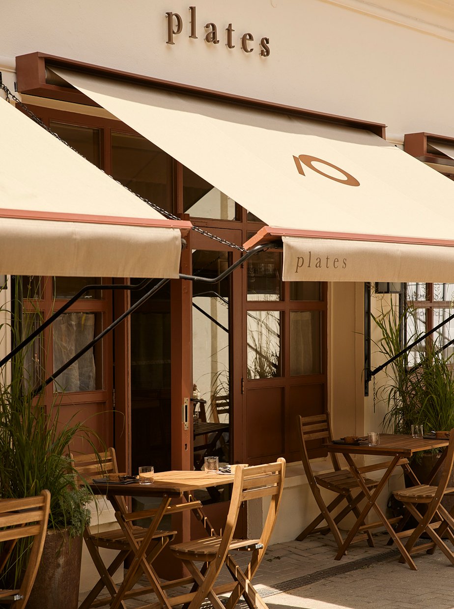 Outdoor seating area of a restaurant named 'plates' with wooden tables and chairs, beige awnings, potted plants, and large windows.