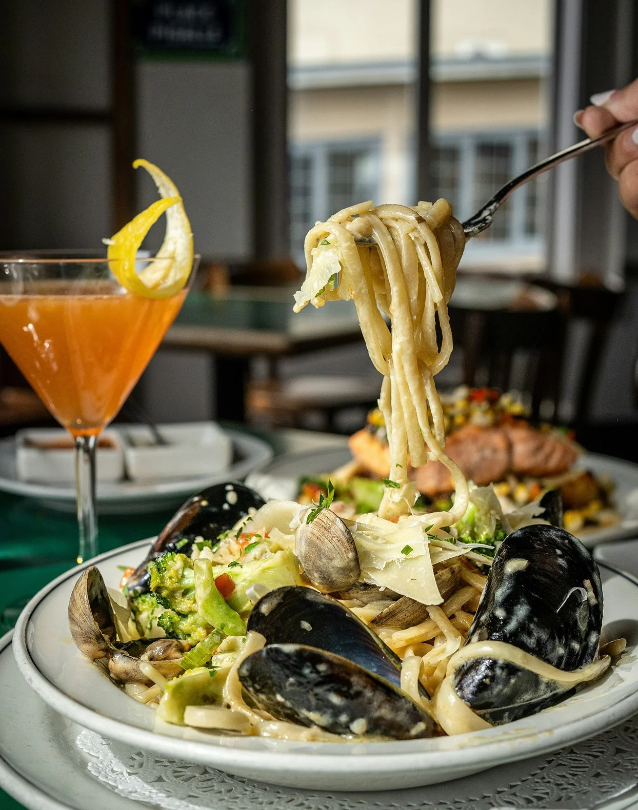 A plate of seafood pasta with mussels, clams, broccoli, and creamy sauce, with a hand lifting a forkful of spaghetti. A cocktail with a lemon twist is in the background.