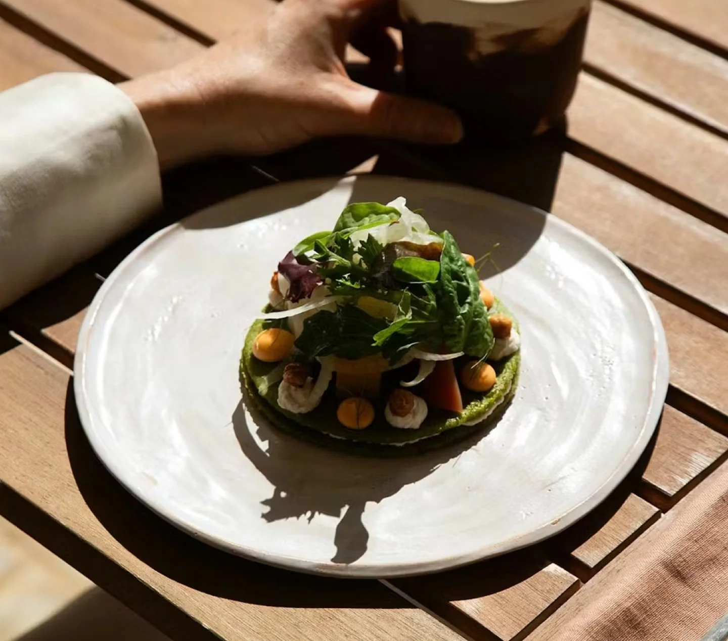 A plate of salad with mixed greens, cherry tomatoes, and other vegetables, served on a white ceramic plate, with a hand holding a cup in the background on a wooden table.