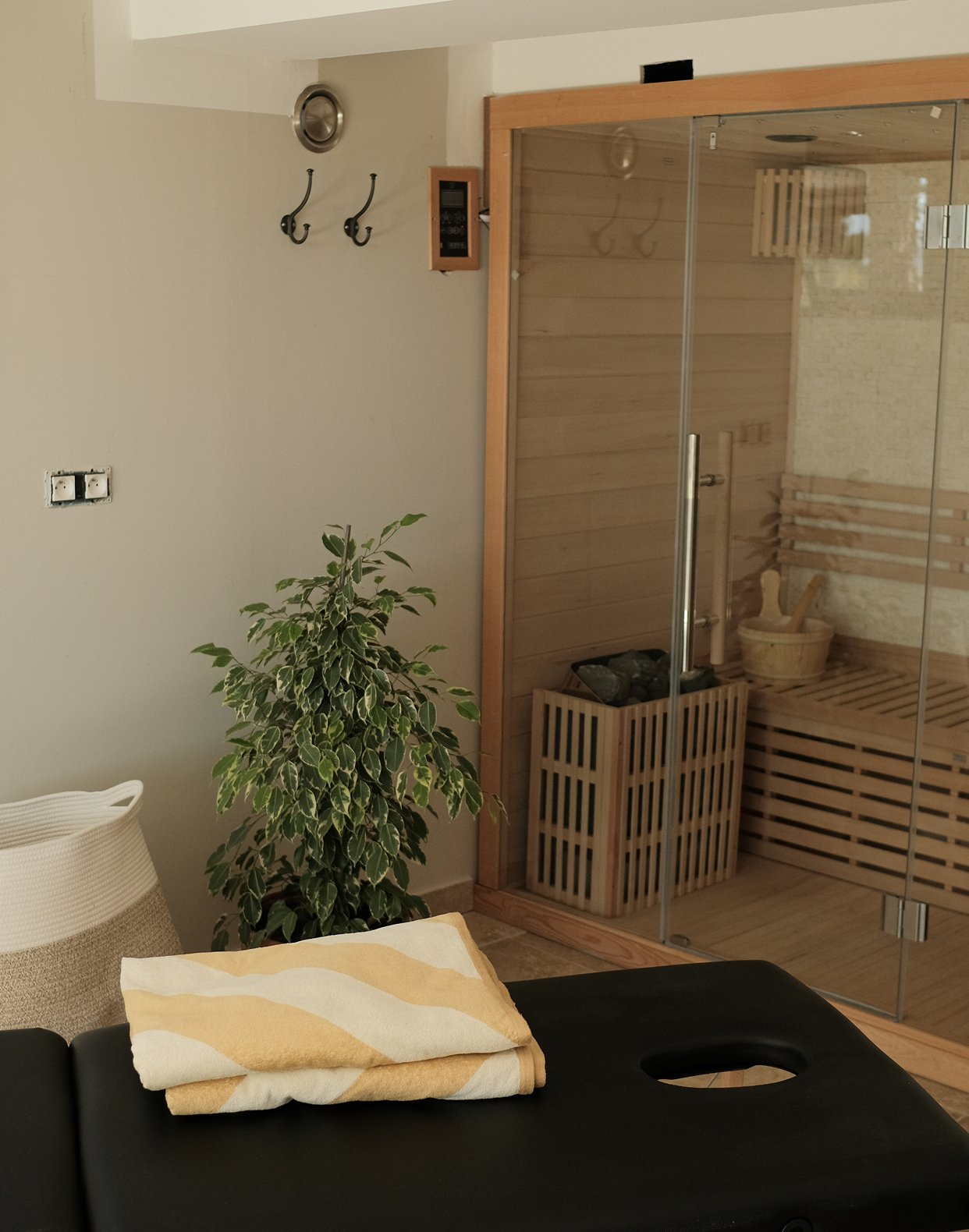 A room with a massage table, towel, and a steam room with a wooden interior, lush green plant, and wall hooks.