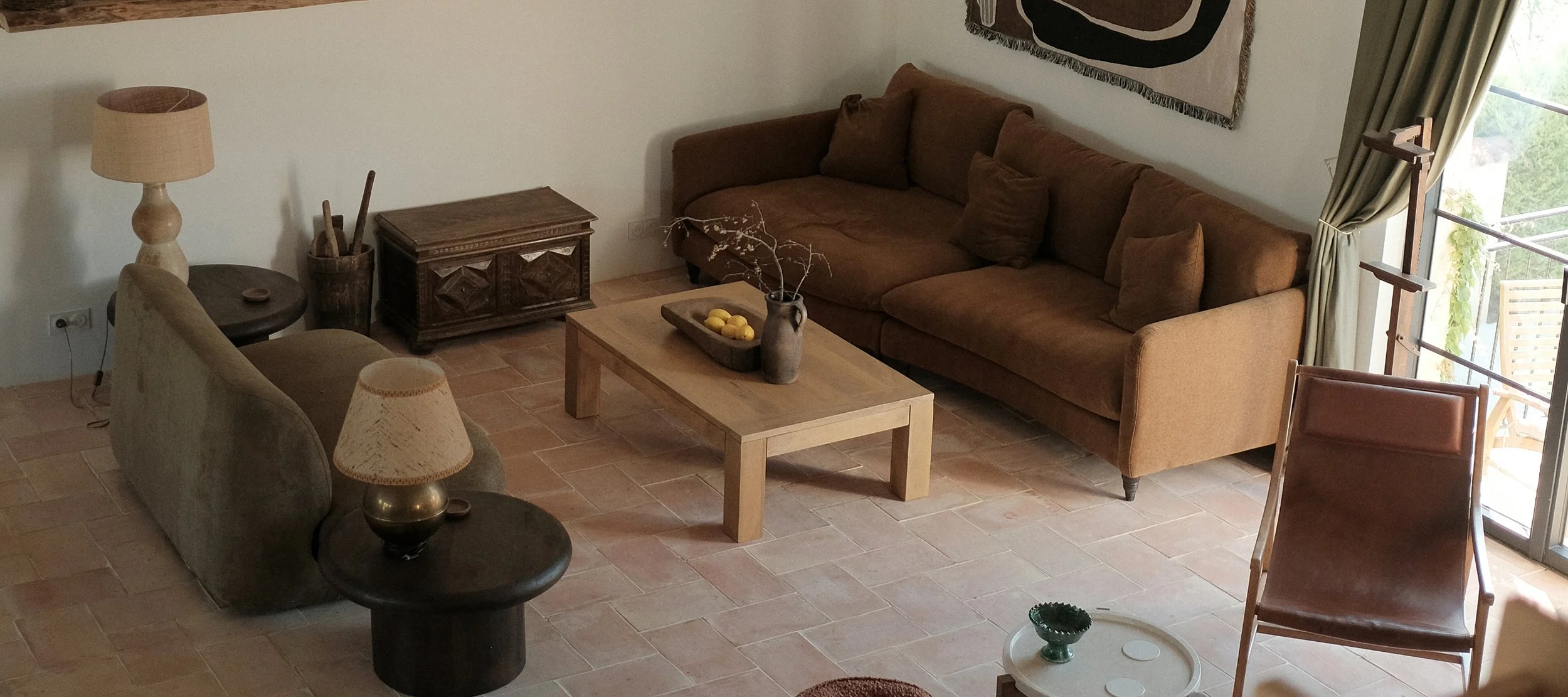 Living room with a brown sofa, a wooden coffee table with a decorative vase and lemons, a dark side table with a lamp, a small wooden cabinet, a beige armchair with a lamp, a balcony door with curtains, and a wall hanging.