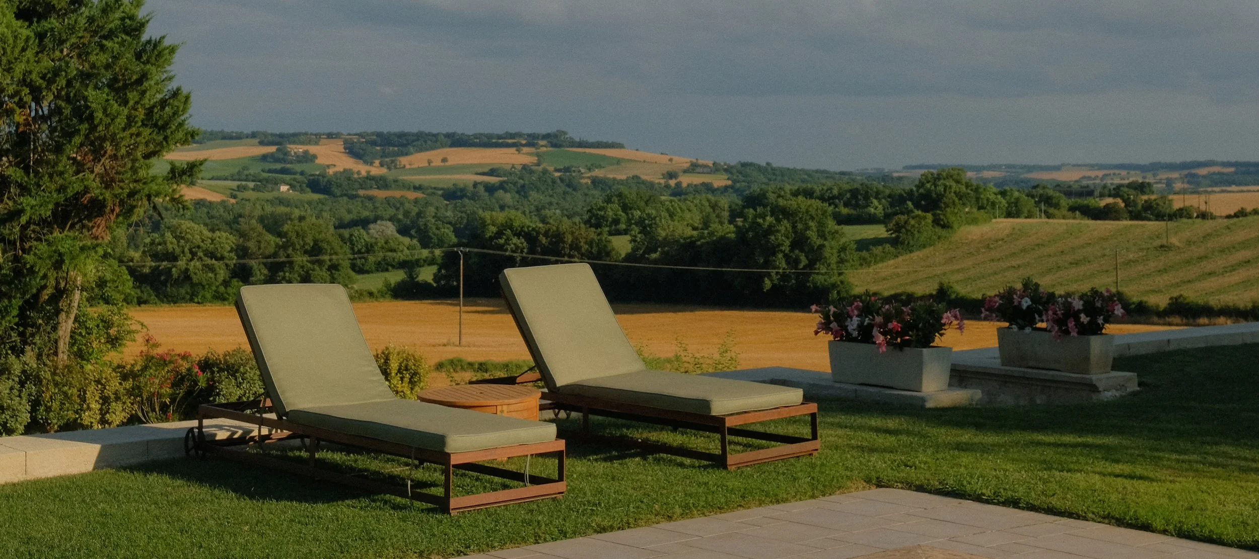 Two outdoor lounge chairs with light cushions on a patio overlooking a scenic landscape of rolling green hills, trees, and farmland under a partly cloudy sky, with two flower pots with pink flowers on the right.