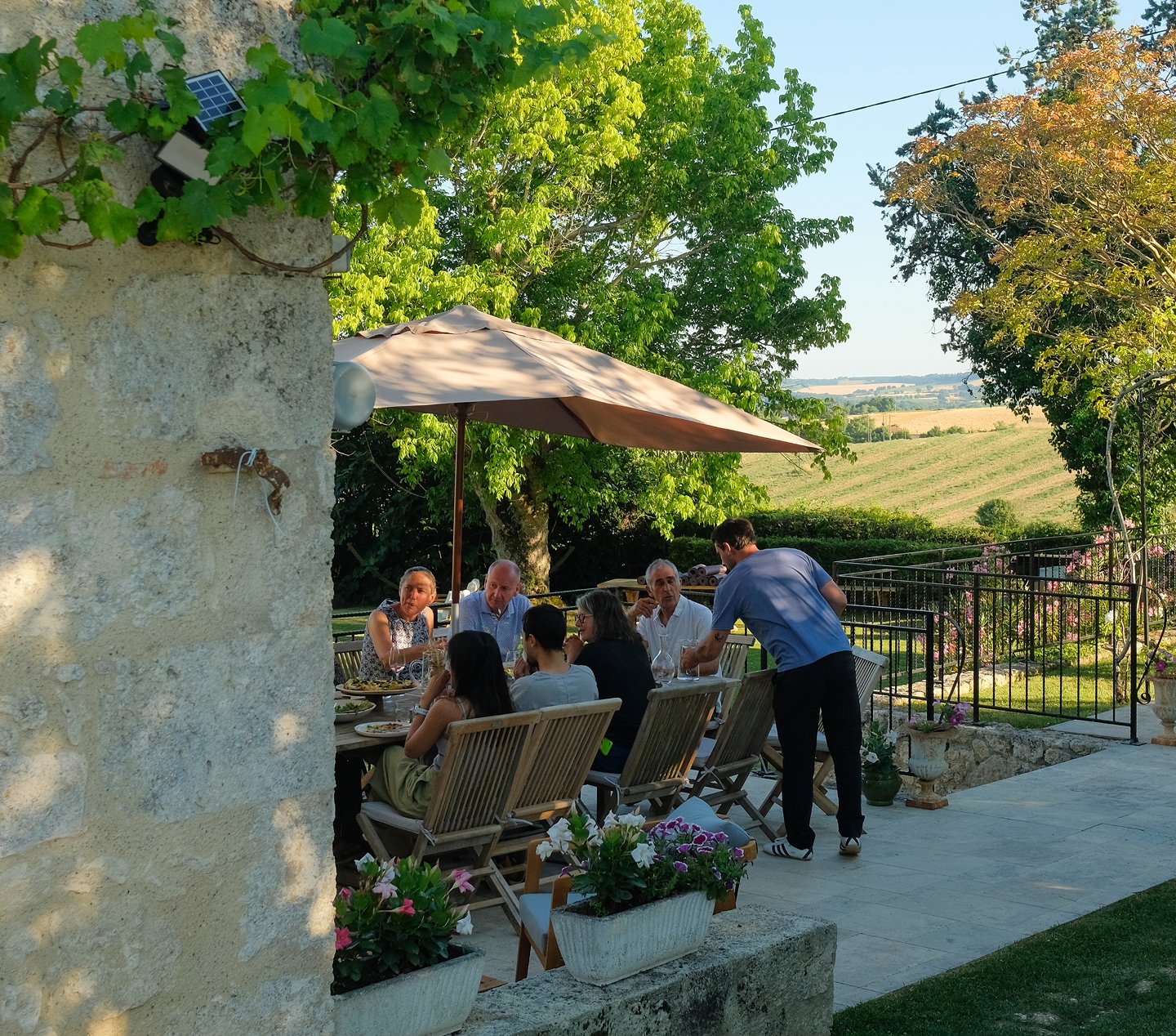 People dining outdoors under a large umbrella on a patio with a scenic view of fields and trees in the background.