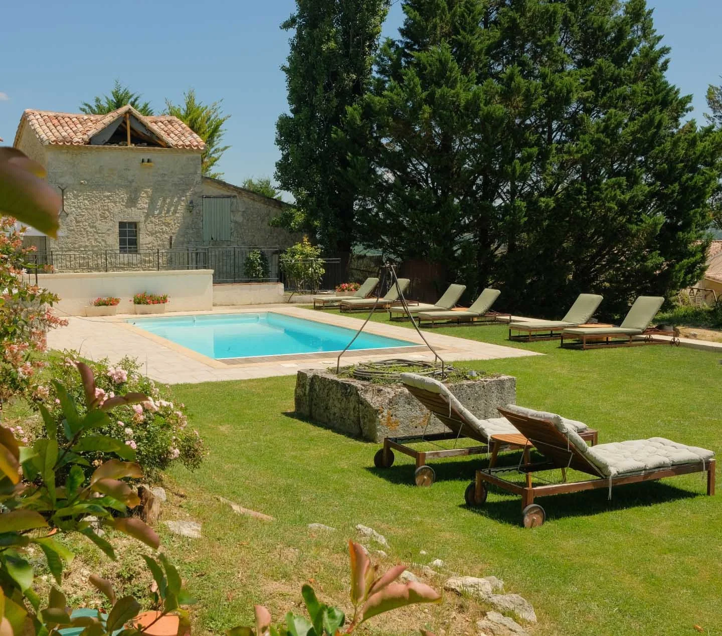 A backyard scene with a rectangular swimming pool surrounded by lounge chairs on a grassy lawn, large trees in the background, and a stone building with a tiled roof.