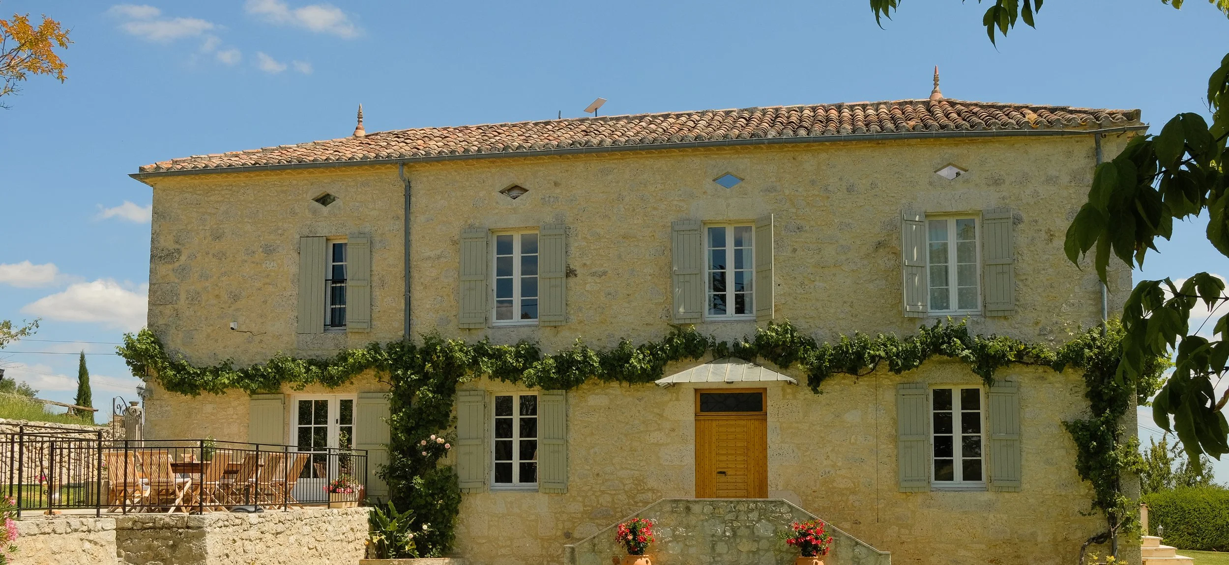 A three-story stone house with green shutters, a tiled roof, and a small balcony area with outdoor furniture, surrounded by greenery and flowering plants, under a blue sky with clouds.