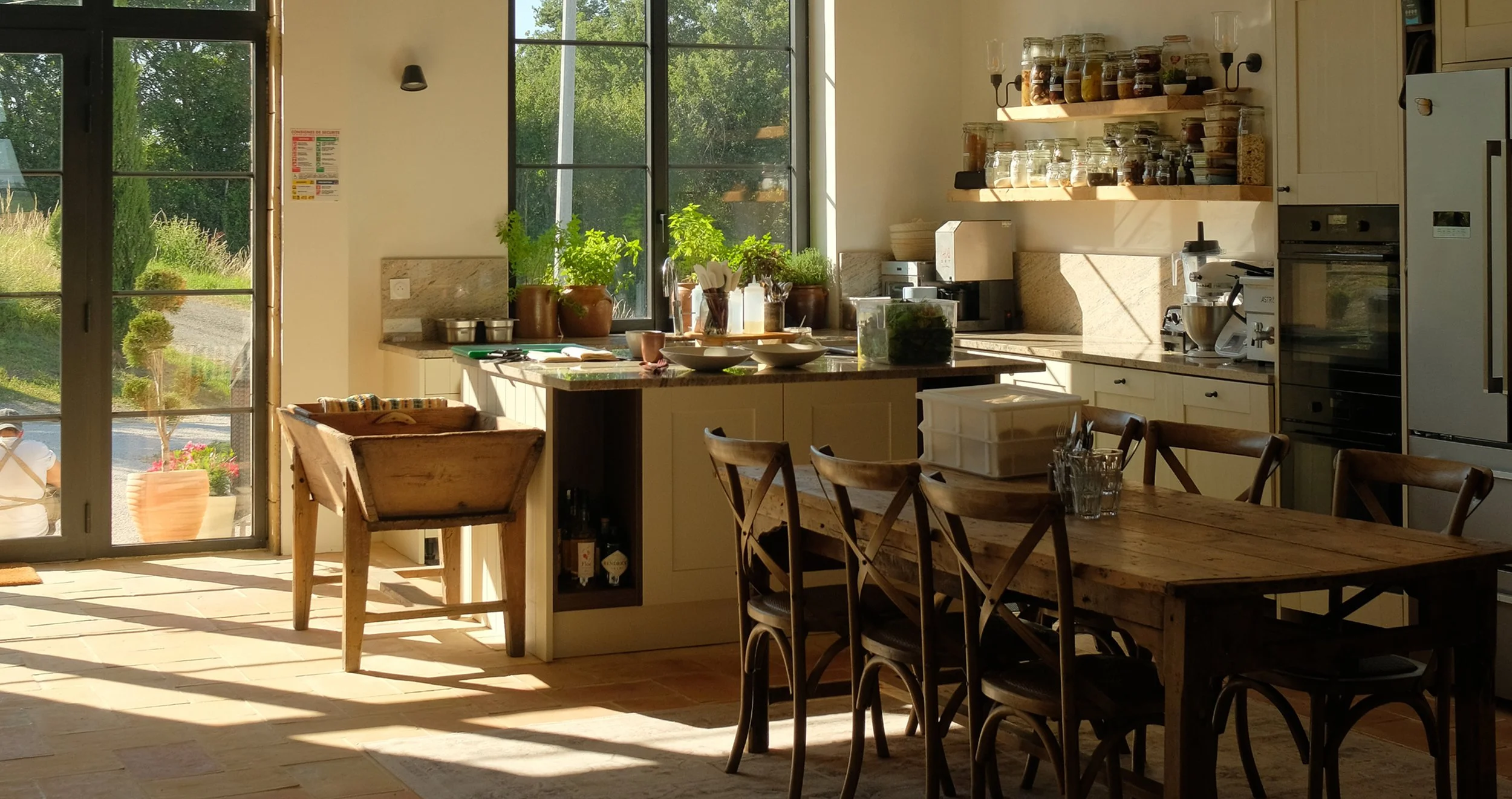 Kitchen with large window, wooden dining table and chairs, kitchen island with bowls and plants, shelves with jars, and sunlight coming through the window.