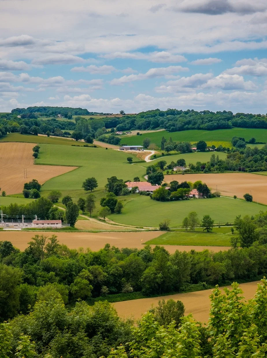Scenic view of rolling countryside with green fields, trees, farmhouses, and a partly cloudy sky.